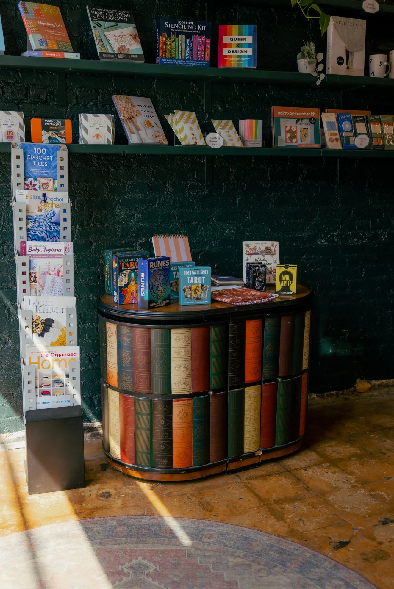 Bookshelves with various craft, art, and lifestyle books, including knitting, crochet, tarot, and home organization, displayed in a cozy bookstore or shop setting.