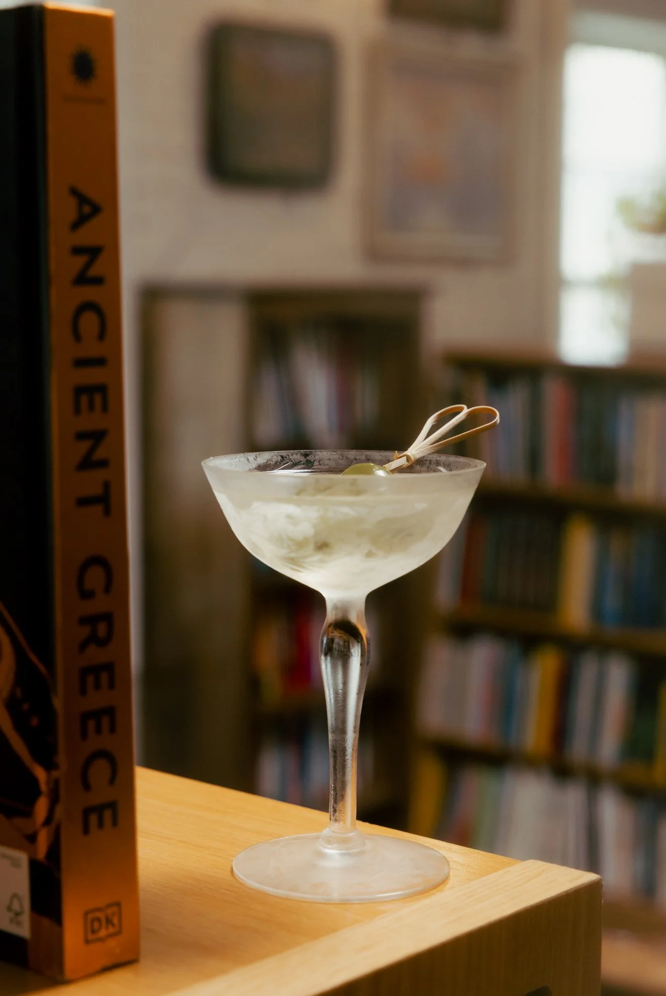 A clear cocktail glass with a stem, filled with a light-colored drink and garnished with a small pick holding green olives. The glass is placed on a wooden surface in a room with bookshelves and framed pictures in the blurred background.