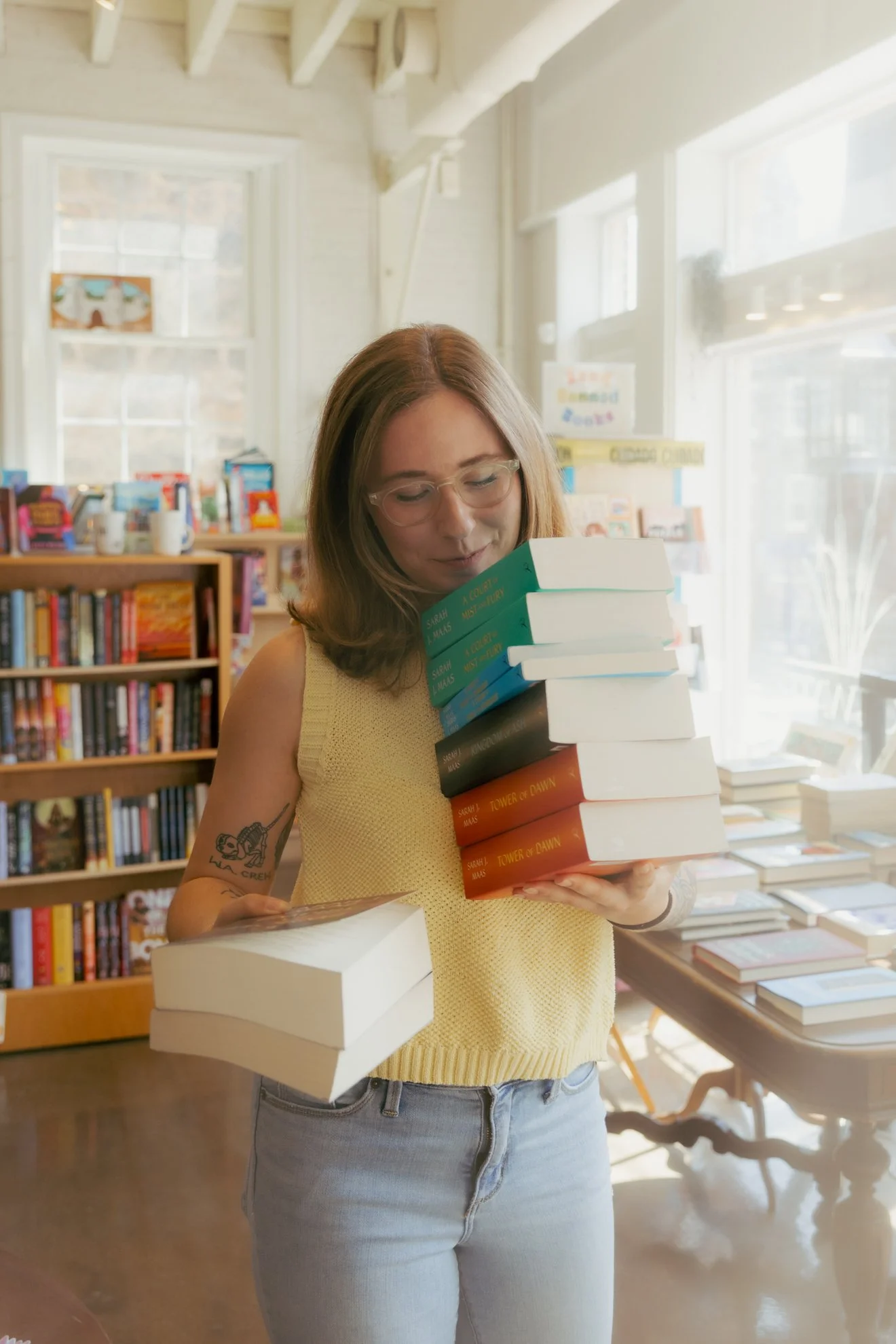 A young woman with glasses, wearing a sleeveless yellow top, holding a stack of books inside a bookstore or library, smiling and looking at the books.