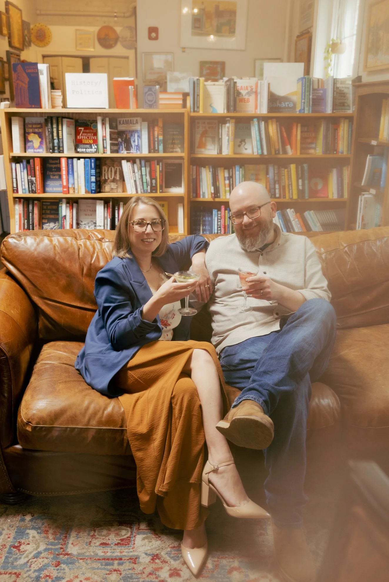 A man and woman sitting closely on a brown leather couch in a library, holding drinks and smiling at the camera. The background features bookshelves filled with books.