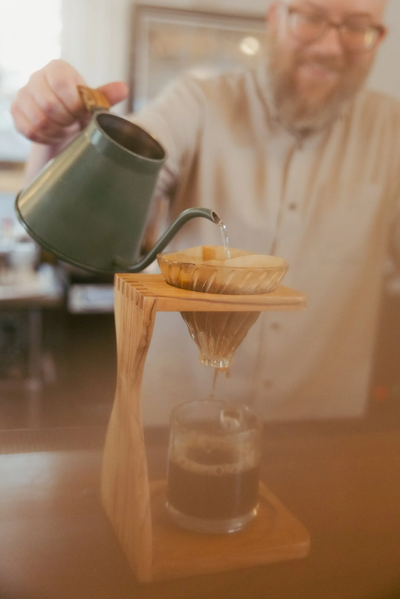 A person pouring liquid from a green watering can into a glass inside a wooden stand with a funnel.
