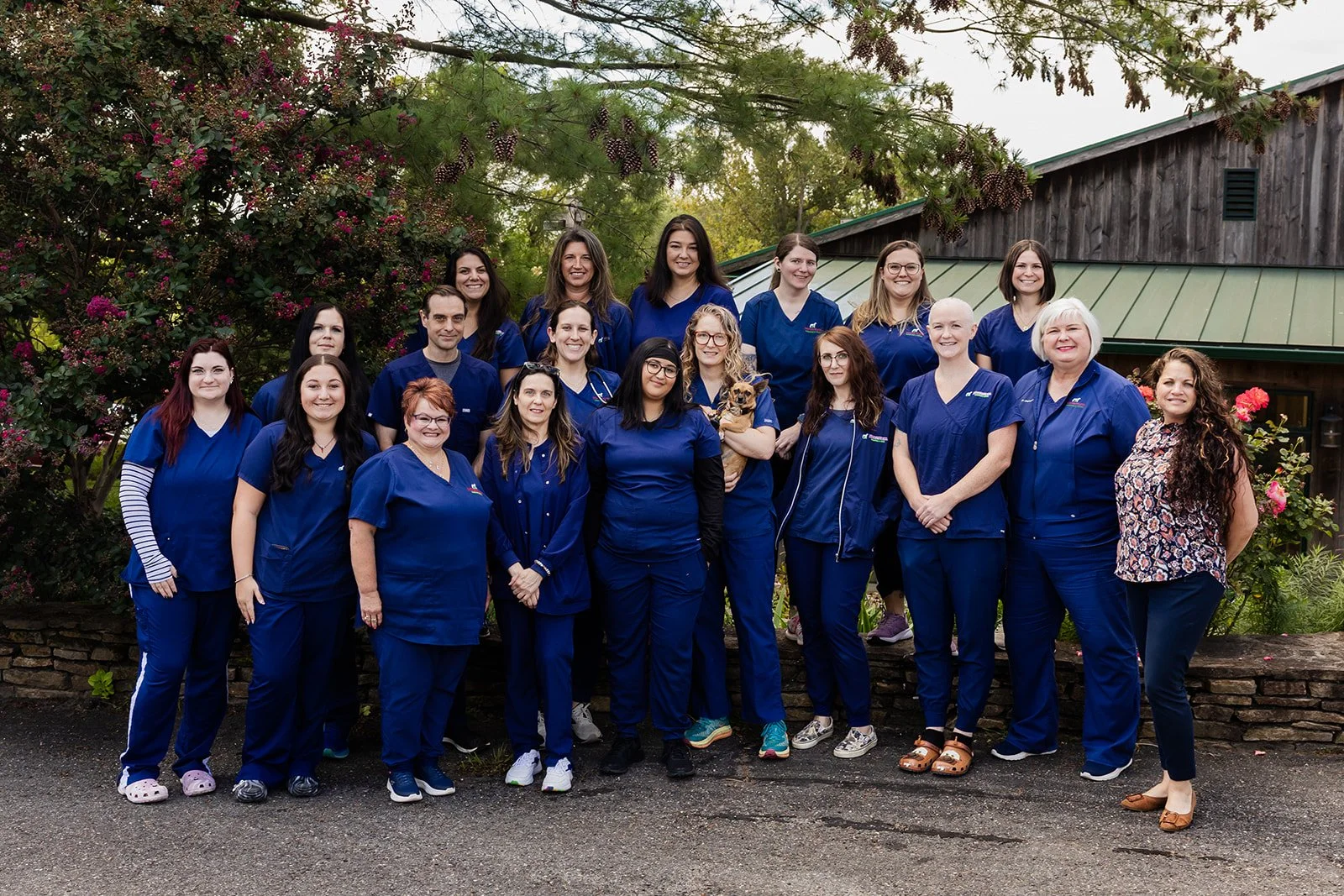 Group of veterinary staff and one dog standing outdoors in front of trees and a building, all wearing blue uniforms.