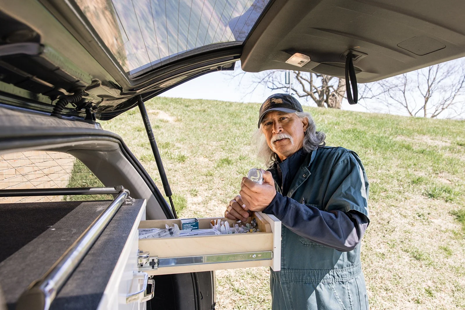 An older man with long gray hair and a mustache is standing outdoors, looking at the camera, holding a pill bottle. He is wearing a black cap and a long-sleeved jacket. There is a drawer of medications with syringes and pill bottles in the open trunk