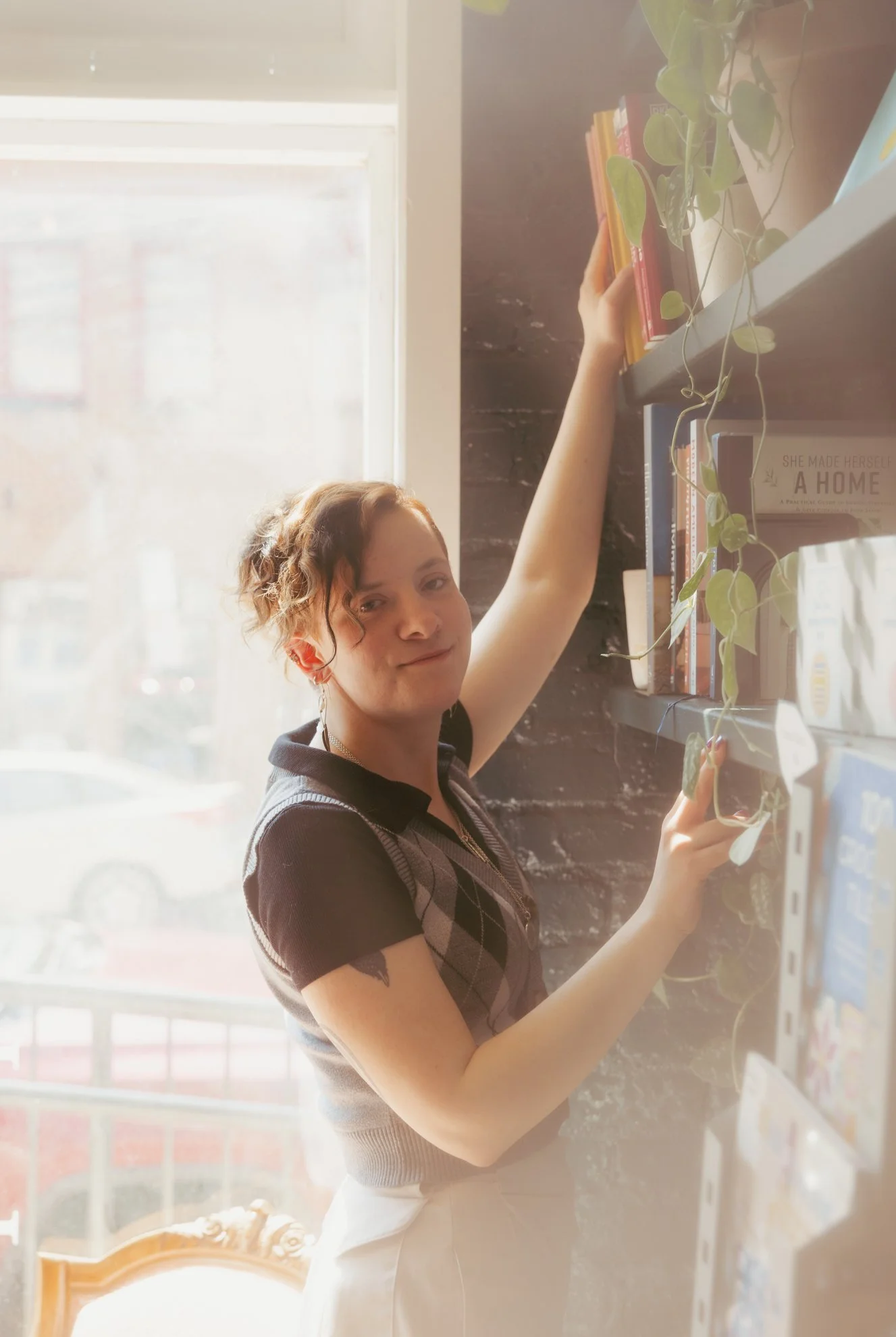 A woman with short hair, wearing a black shirt with argyle pattern, is reaching for a book on a shelf in a cozy space with a large window and a green plant nearby.