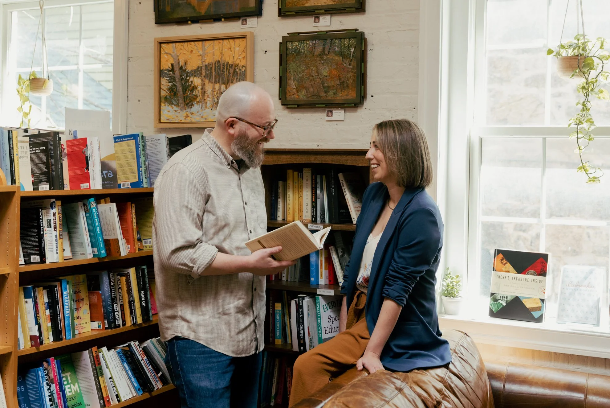 A man with glasses and a beard holding a book is smiling and talking to a woman with shoulder-length hair, who is sitting on a leather tufted bench. They are in a bookstore or library with shelves of books and some artwork on the brick wall behind th