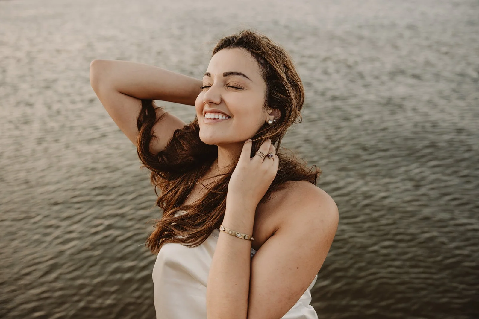 A happy woman with long, wavy brown hair posing near a body of water with her eyes closed and a smile on her face, wearing jewelry and a white top.