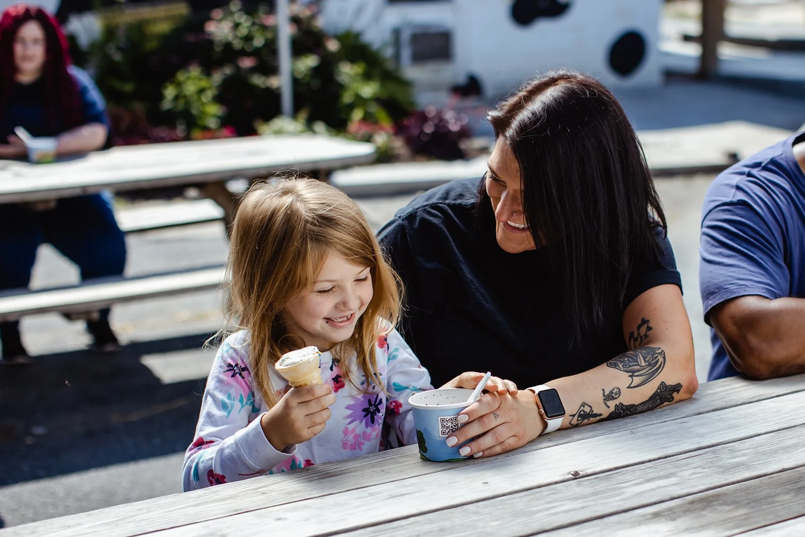 A woman and a young girl share a moment at a picnic table, smiling and enjoying ice cream together outdoors. In the background, another person is out of focus sitting at a separate table.