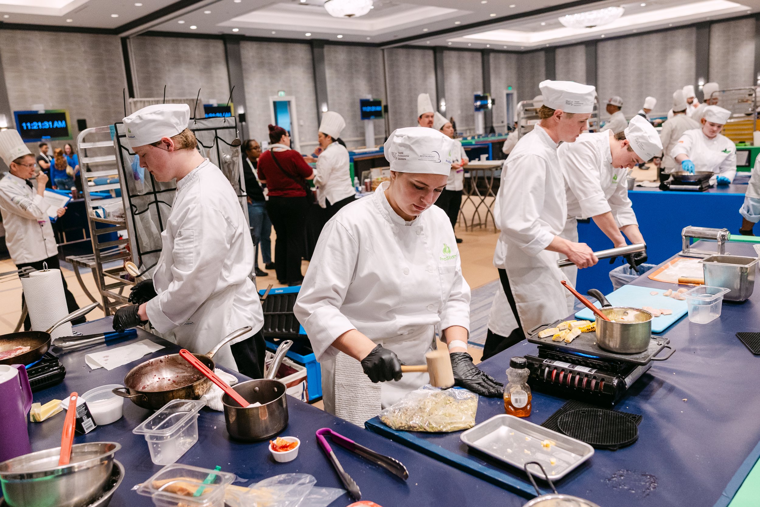 Group of people dressed as chefs working in a professional kitchen during a cooking class or competition in a large conference room.