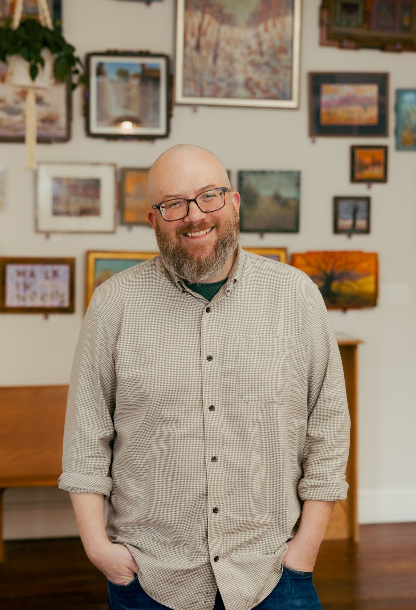 A smiling man with glasses and a beard, standing in front of a wall with multiple framed paintings, wearing a beige checkered shirt and jeans.