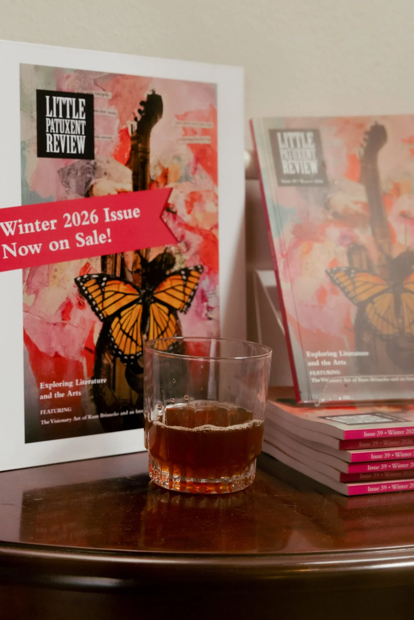 A printed magazine or brochure titled 'LITTLE PATUXENT REVIEW' is displayed on a table, with a red banner indicating 'Winter 2026 Issue Now on Sale!'. The cover features a black silhouette of a guitar neck, a colorful watercolor background, a butterf