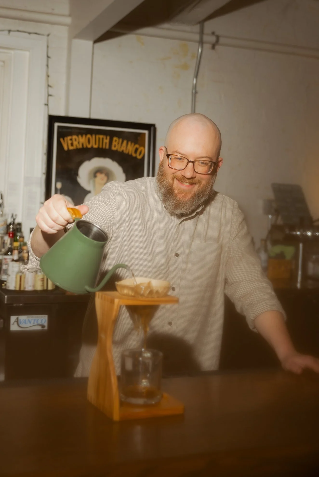 A man with glasses and a beard pouring coffee into a glass on a wooden coaster in a cozy setting. In the background, there is a framed vintage poster with yellow text that reads 'VERMOUTH BIANCO' and a partially visible bar area.