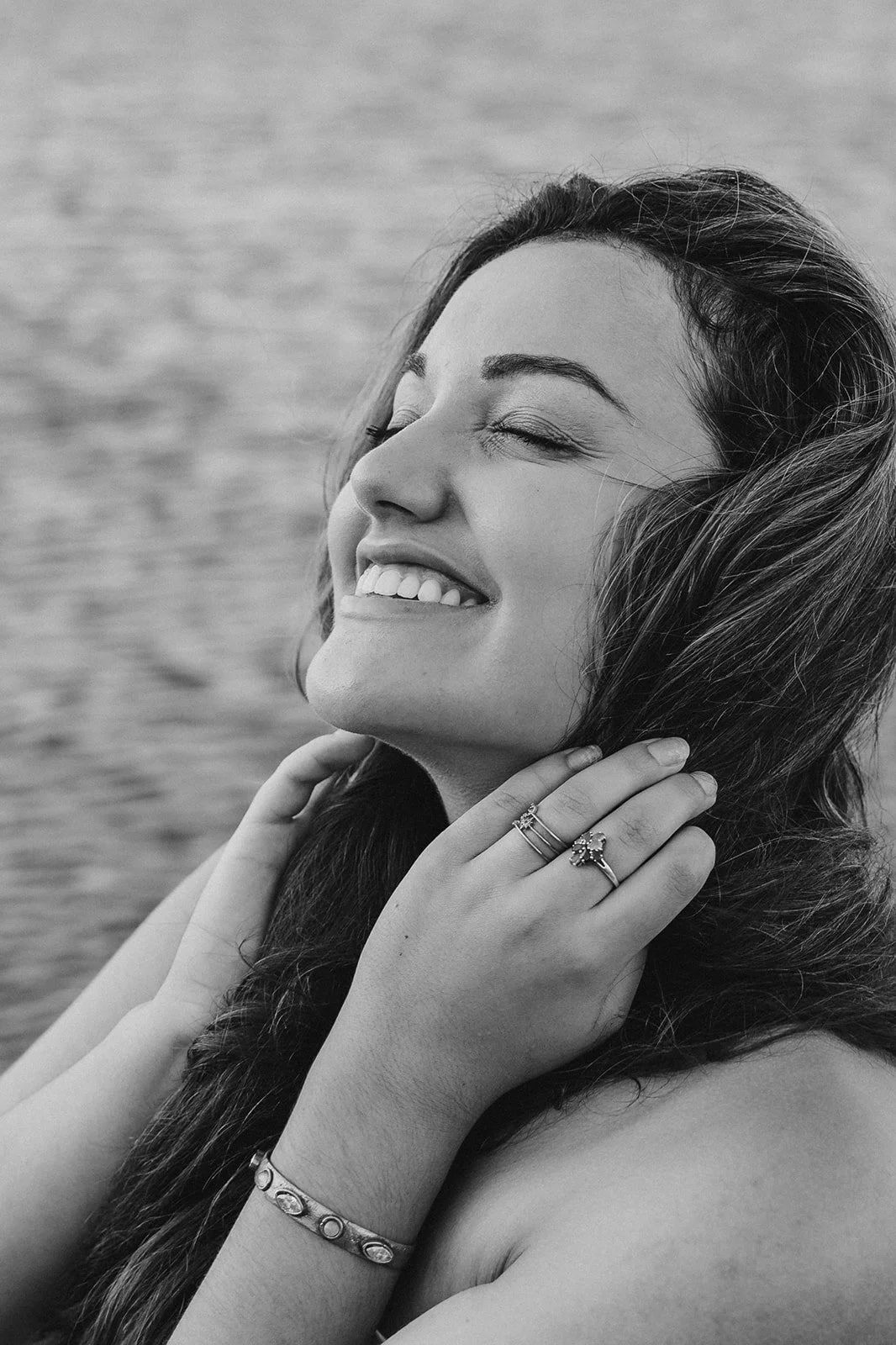 A black and white photo of a woman smiling with her eyes closed, touching her neck with her left hand, wearing jewelry, with water in the background.