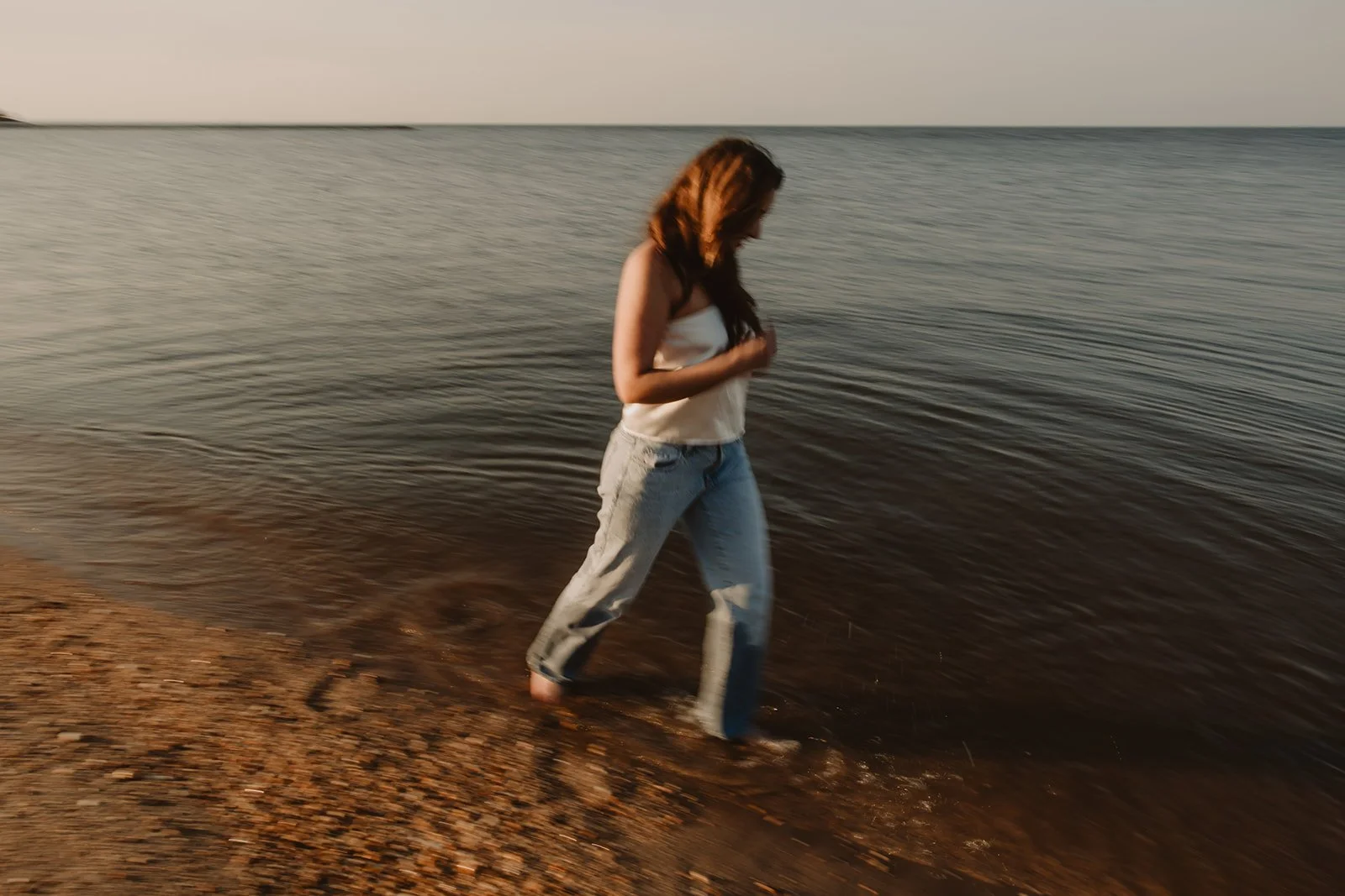 A woman with long brown hair, wearing a white tank top and light blue jeans, wading in the shallow water at the beach during sunset.