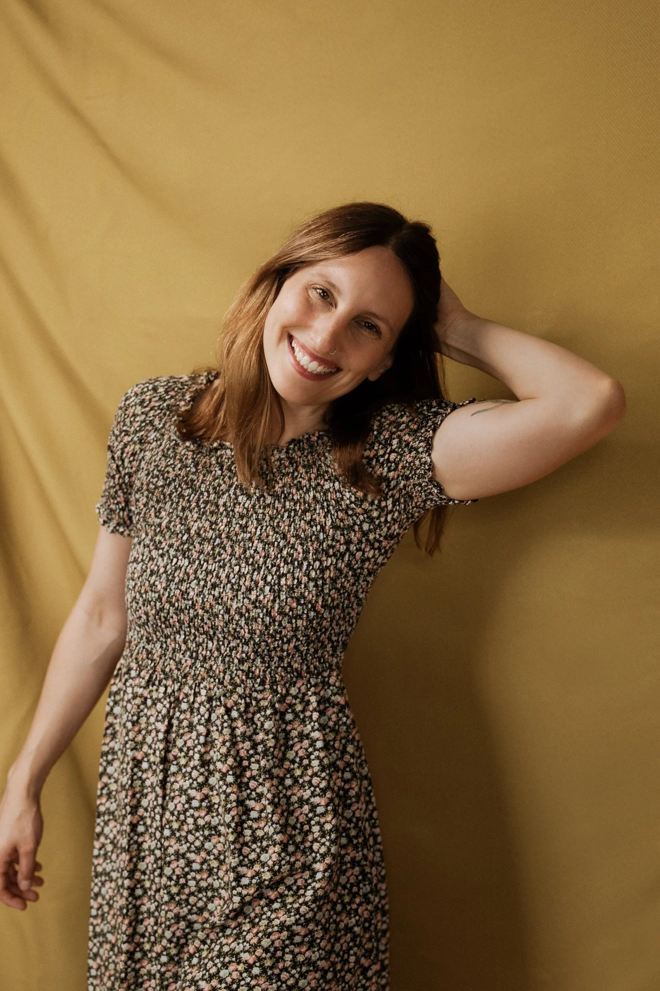 A woman with long brown hair, wearing a floral dress, smiling and leaning against a yellow wall with her left arm behind her head.