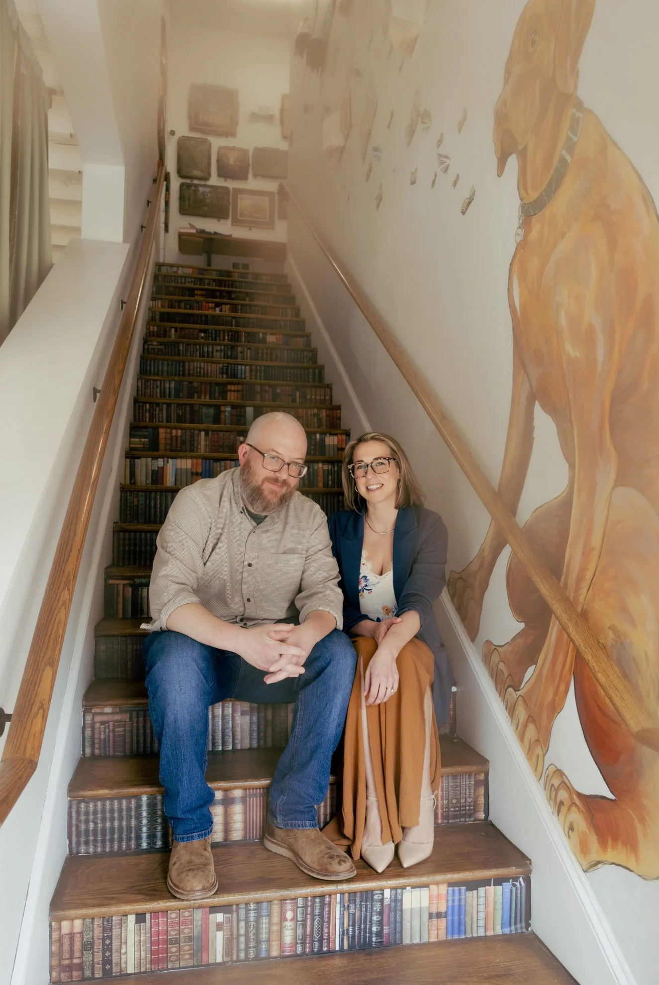 A man and woman sitting on a staircase with a mural of a large dog on the wall beside them. The staircase is decorated with a design resembling books, and there are framed pictures hanging on the wall at the top of the stairs.