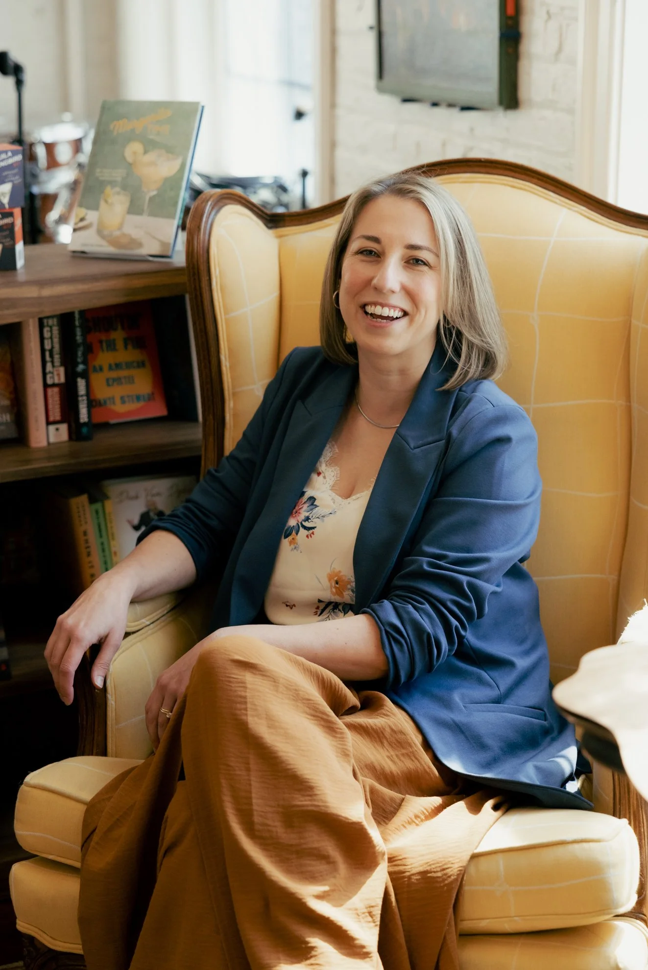 A woman with shoulder-length gray hair smiling, sitting on a yellow armchair in a cozy room with books and a TV in the background.