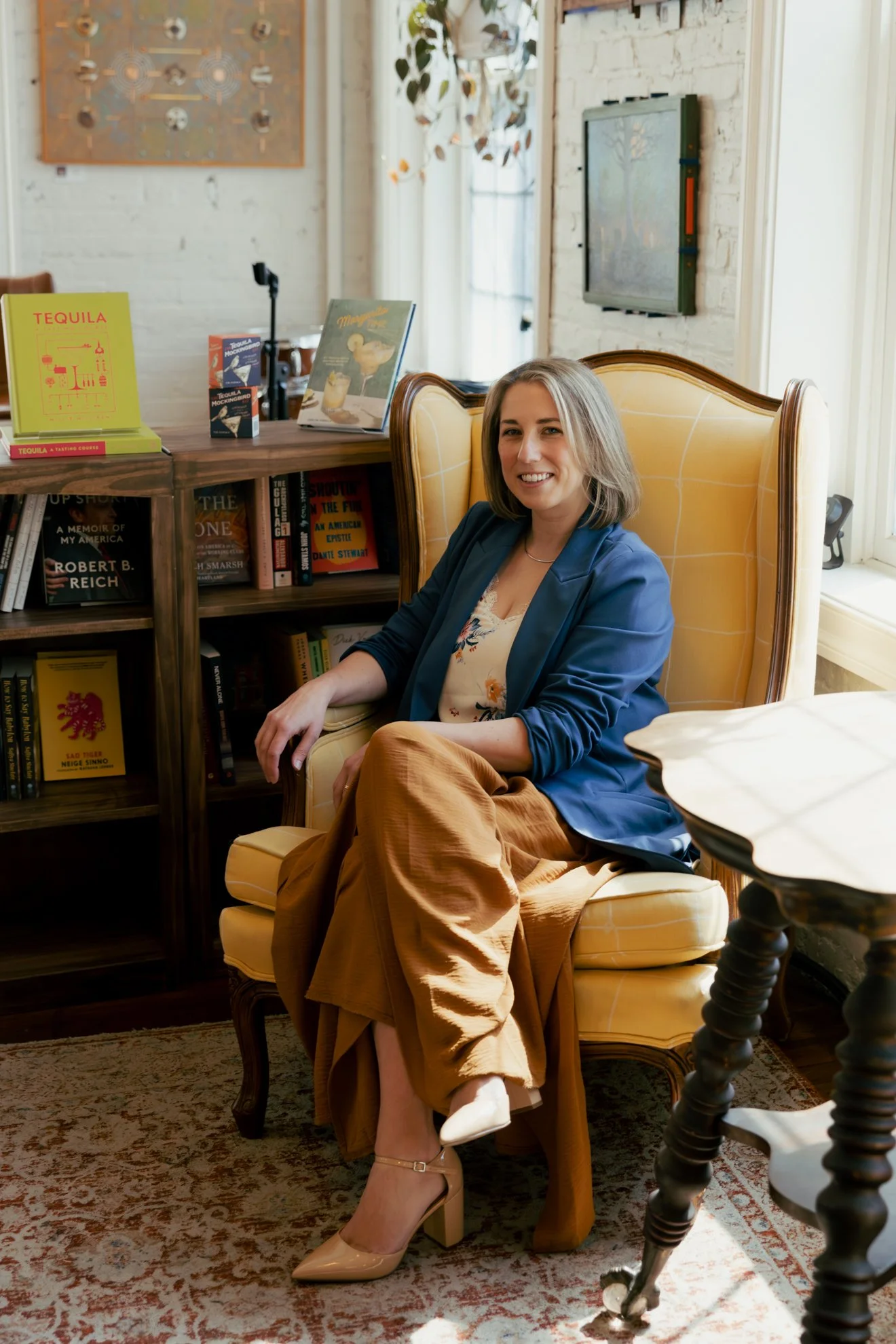 A woman with shoulder-length blonde hair sitting in a yellow armchair in a cozy, well-lit room with white brick walls. She is smiling and dressed in a blue blazer, floral blouse, and tan wide-leg pants, wearing beige high heels. Behind her is a books