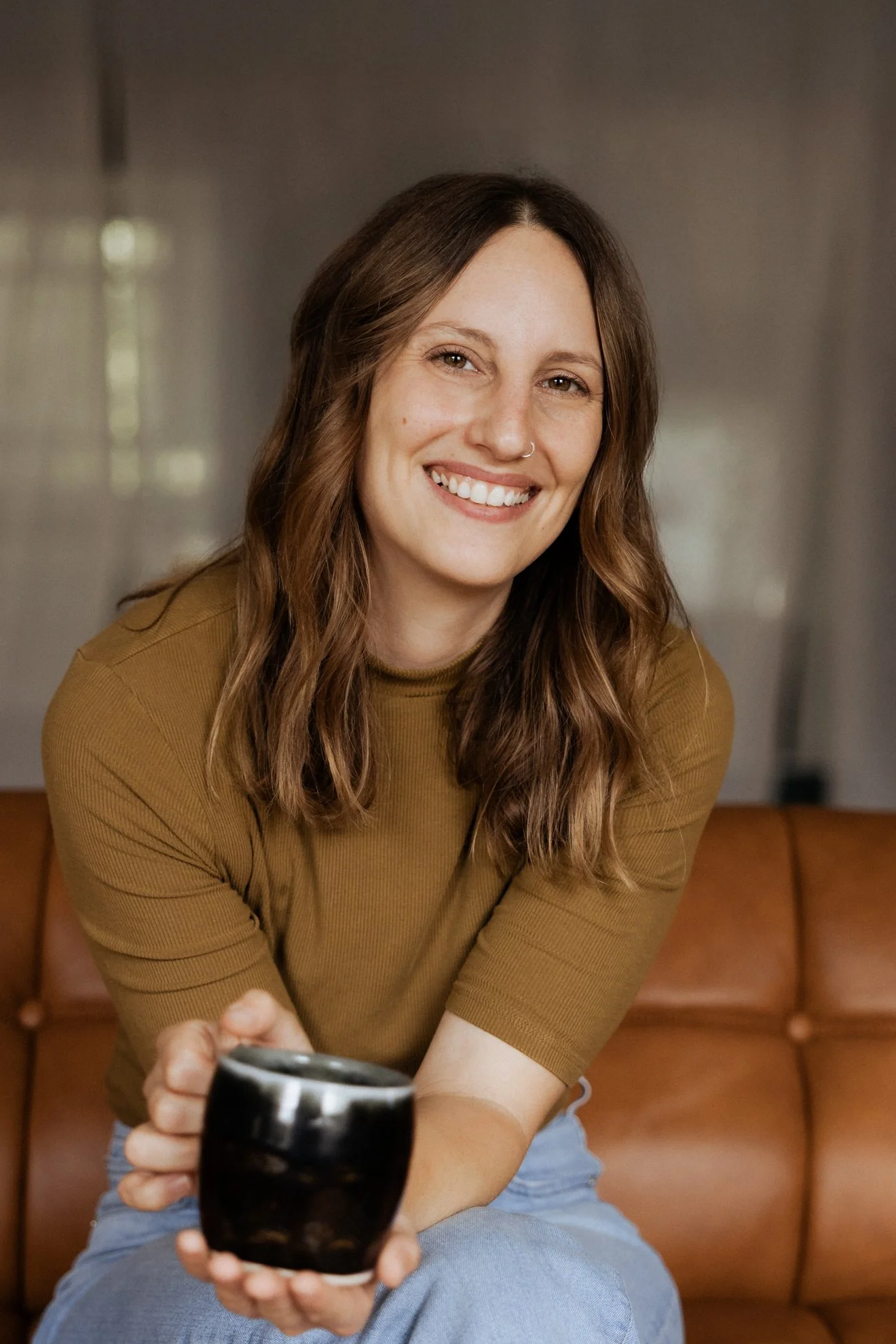 Smiling young woman with curly brown hair and a nose ring, sitting on a brown leather couch, holding a black mug, wearing a mustard-colored top and light blue jeans, in a room with beige walls.