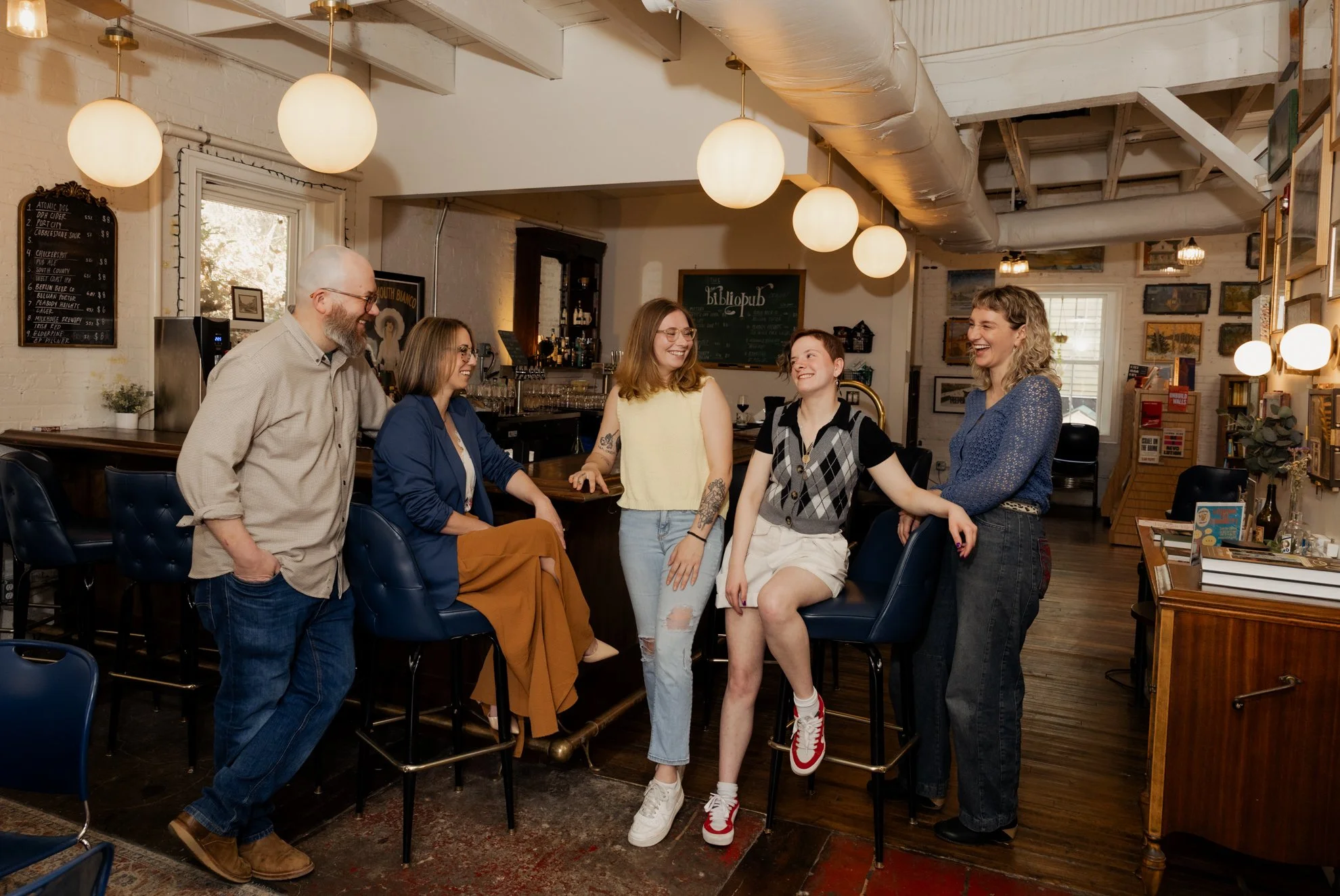 Seven people, four women and three men, are smiling and laughing together in a cozy cafe with white walls, wooden furniture, and warm lighting.