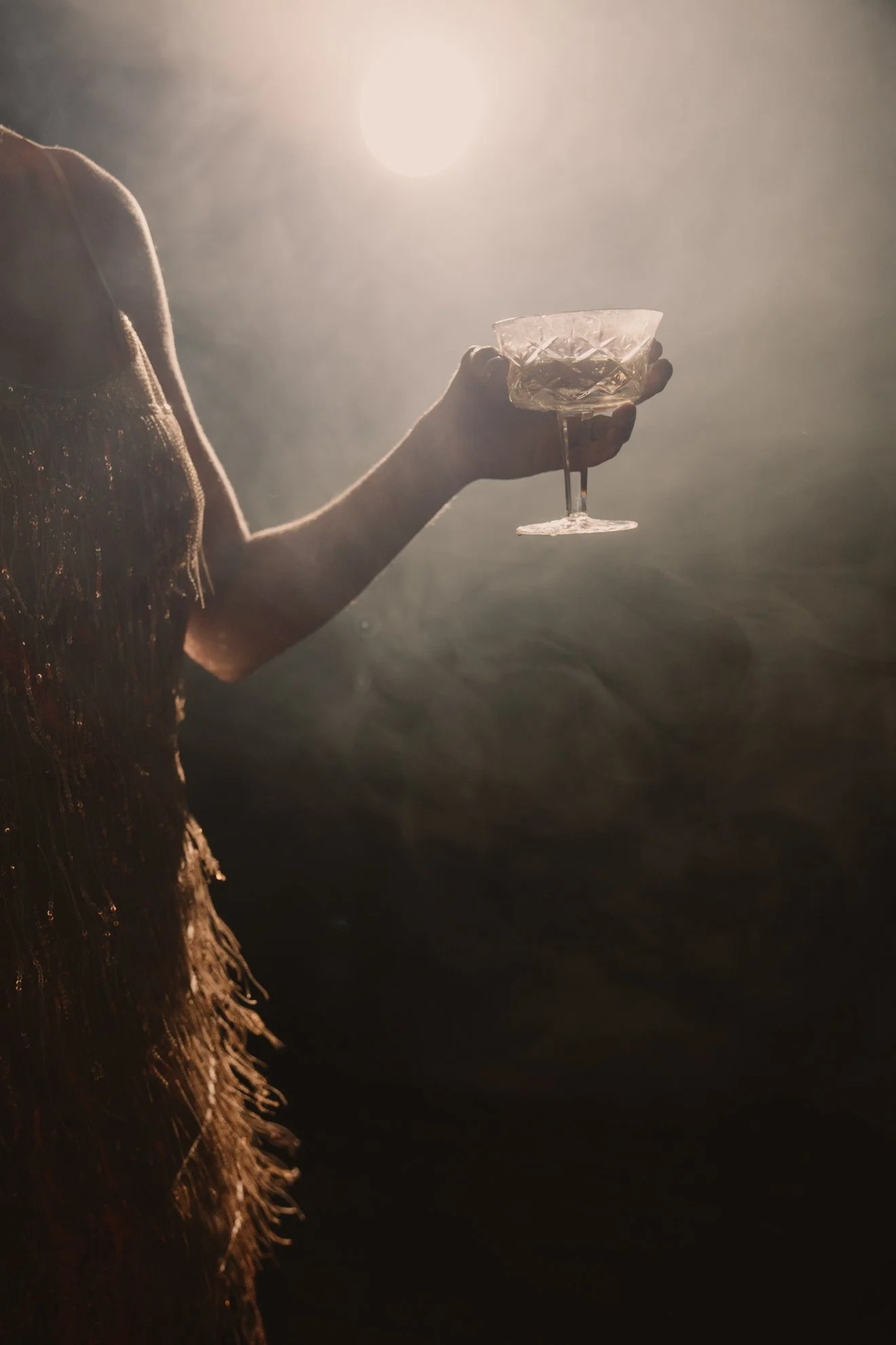 A person wearing a sparkly dress holding a glass of champagne or white wine, with the sun in the background, creating a backlit scene.