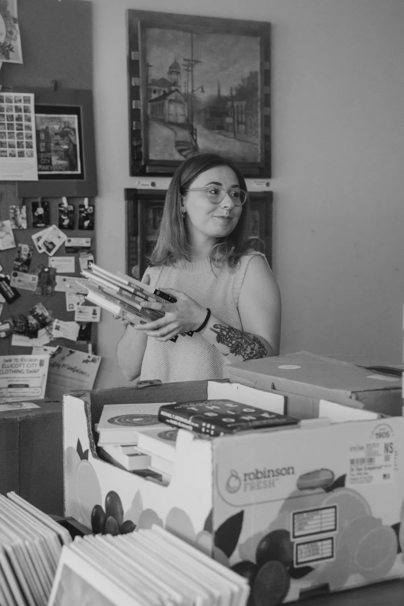 A woman with glasses, shoulder-length hair, and a tattoo on her left arm, holding a stack of books in a store or library. Behind her are shelves filled with books and a bulletin board with various papers and notices. Decorative paintings or posters a