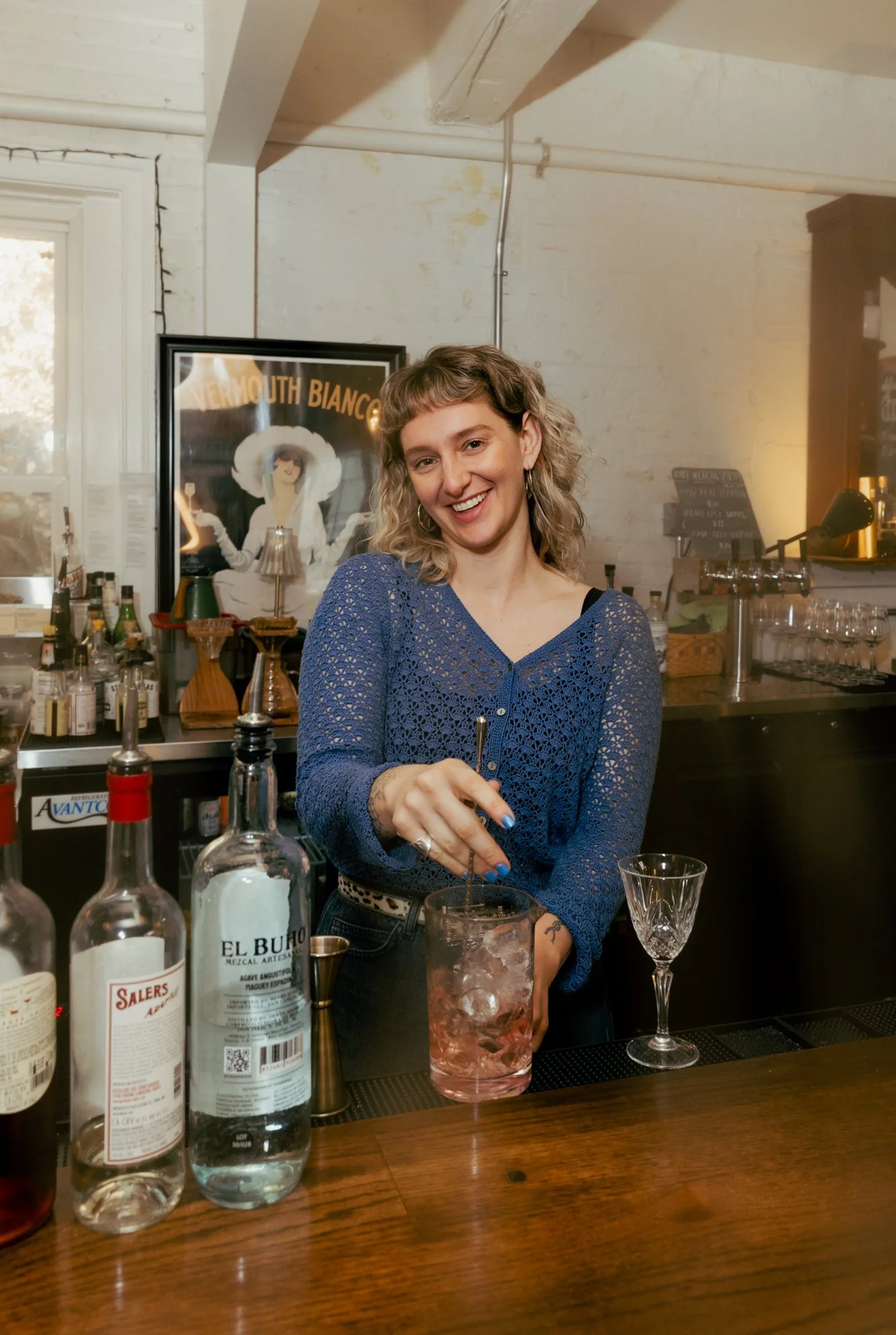 A woman with curly blonde hair smiling while preparing a drink behind a bar with liquor bottles, glasses, and bar tools.