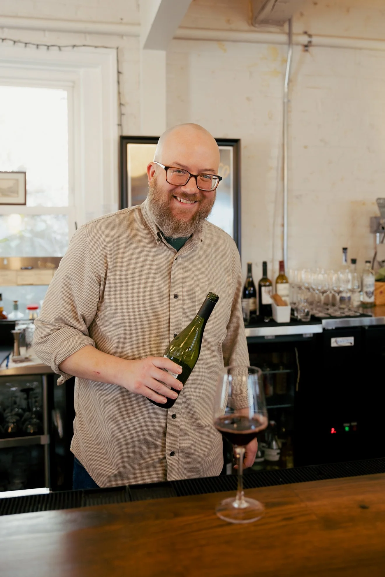 A smiling man with a bald head and beard, wearing glasses and a checked shirt, holding a bottle of wine and a glass of wine in a bar or restaurant setting.