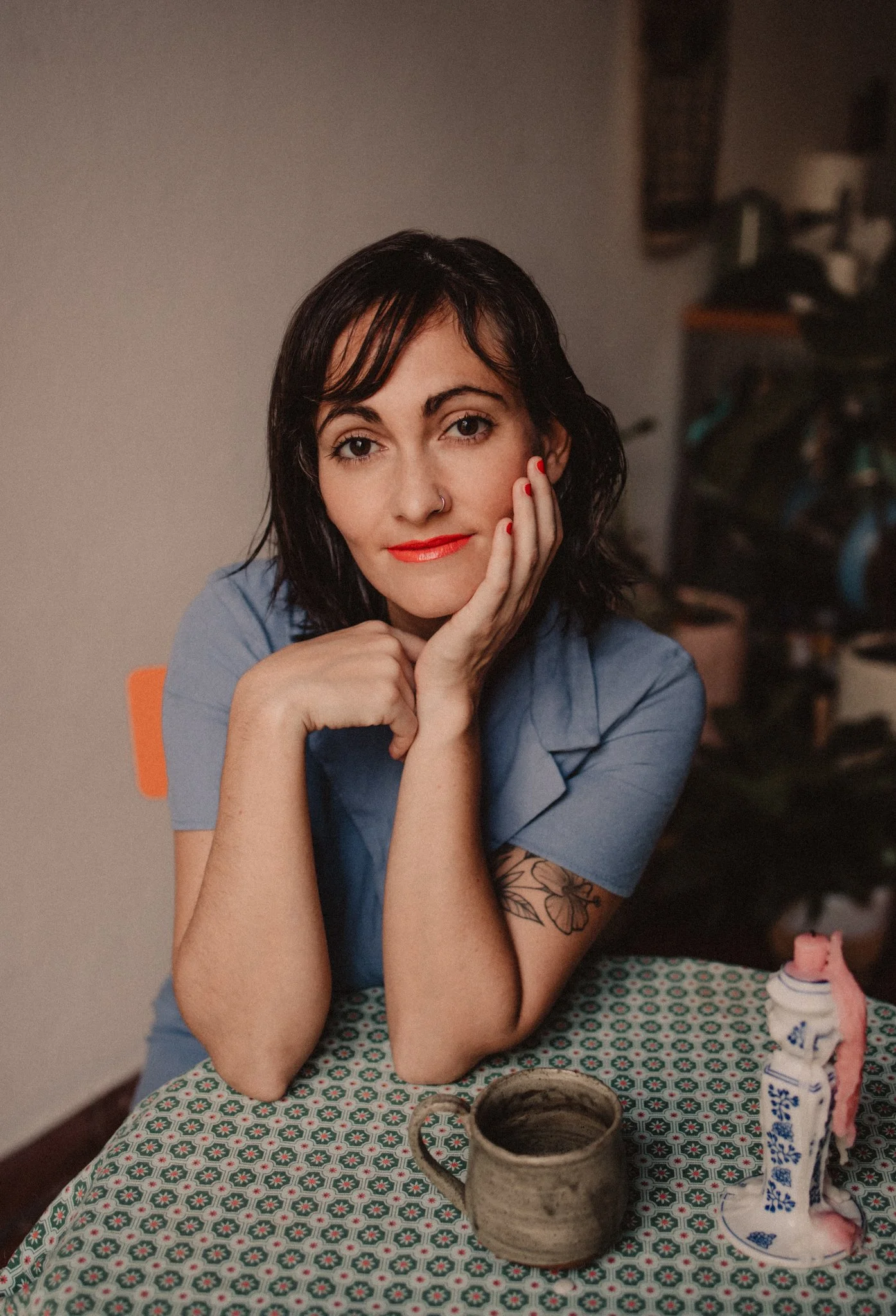 A woman with dark, wavy hair and red lipstick, sitting at a table with a patterned tablecloth, resting her chin on her hand and looking at the camera. There are a mug and a decorative bottle on the table.