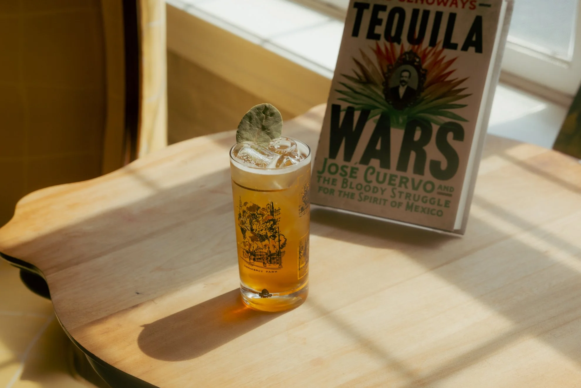 A glass of iced beverage garnished with a green leaf placed at the edge of the glass, with a book titled 'Tequila Wars' standing upright on a wooden surface near the glass.