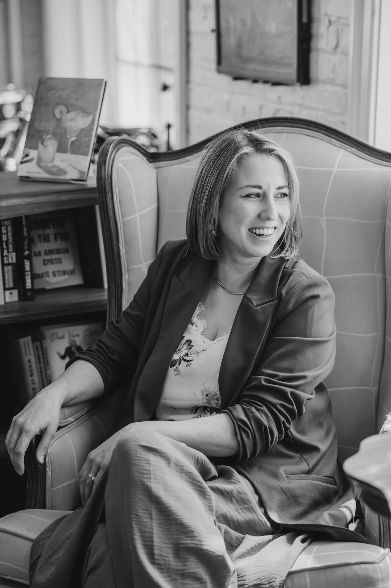 A woman sitting on a cushioned armchair, smiling and looking to her left, with a bookshelf and television in the background.