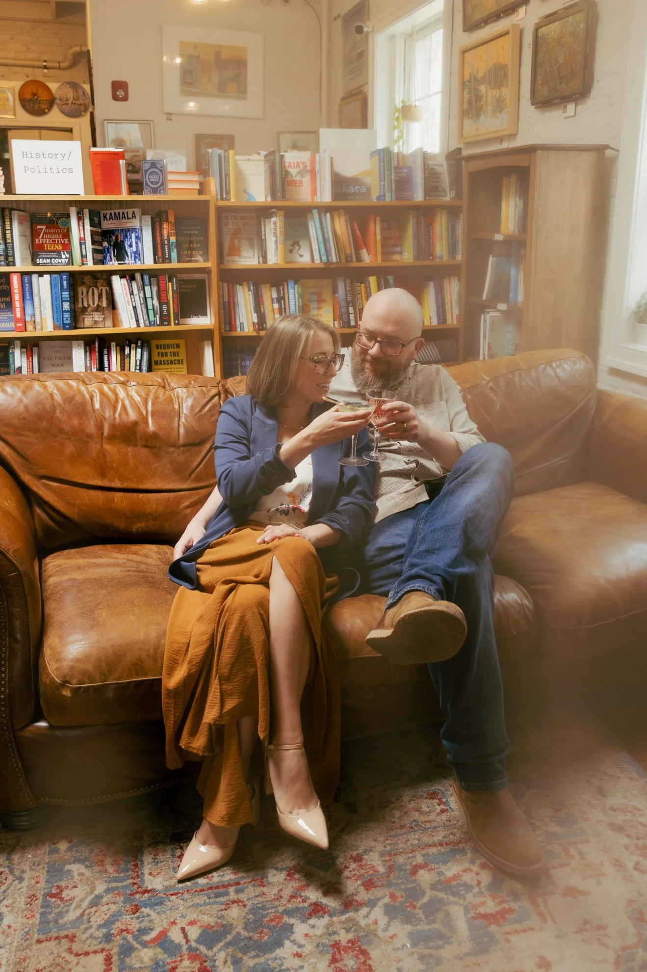 A man and woman sitting on a leather couch inside a library or bookstore, sharing a moment as they toast with drinks. The woman is wearing a blue jacket, a floral skirt, and cream-colored heels, while the man is bald, has a beard, and is wearing glas