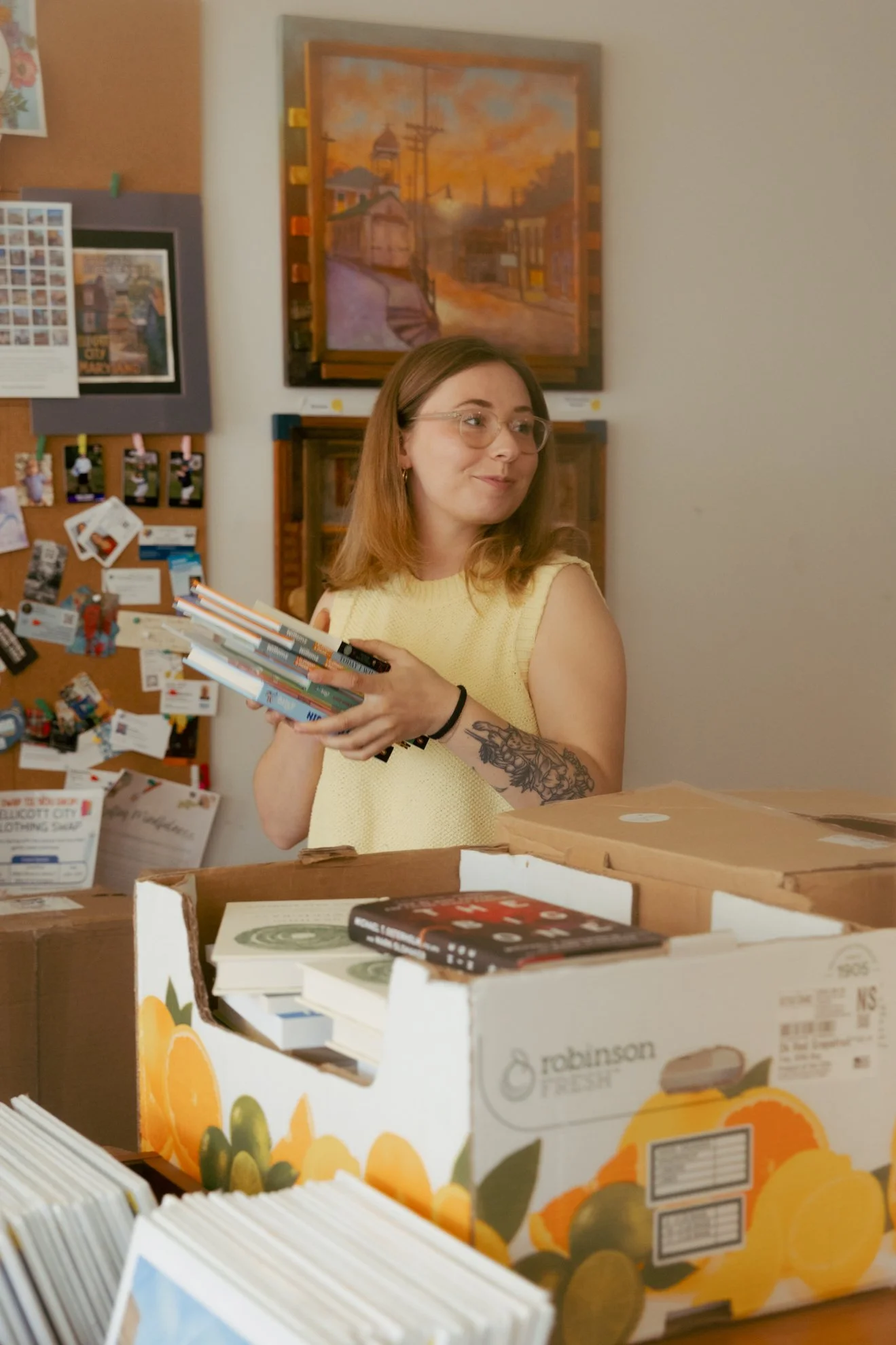 Young woman with glasses, shoulder-length brown hair, and a sleeve tattoo on her left arm, holding books in a room with framed artwork and papers on the wall. The room has a table with stacked books and a box with a lemon design.