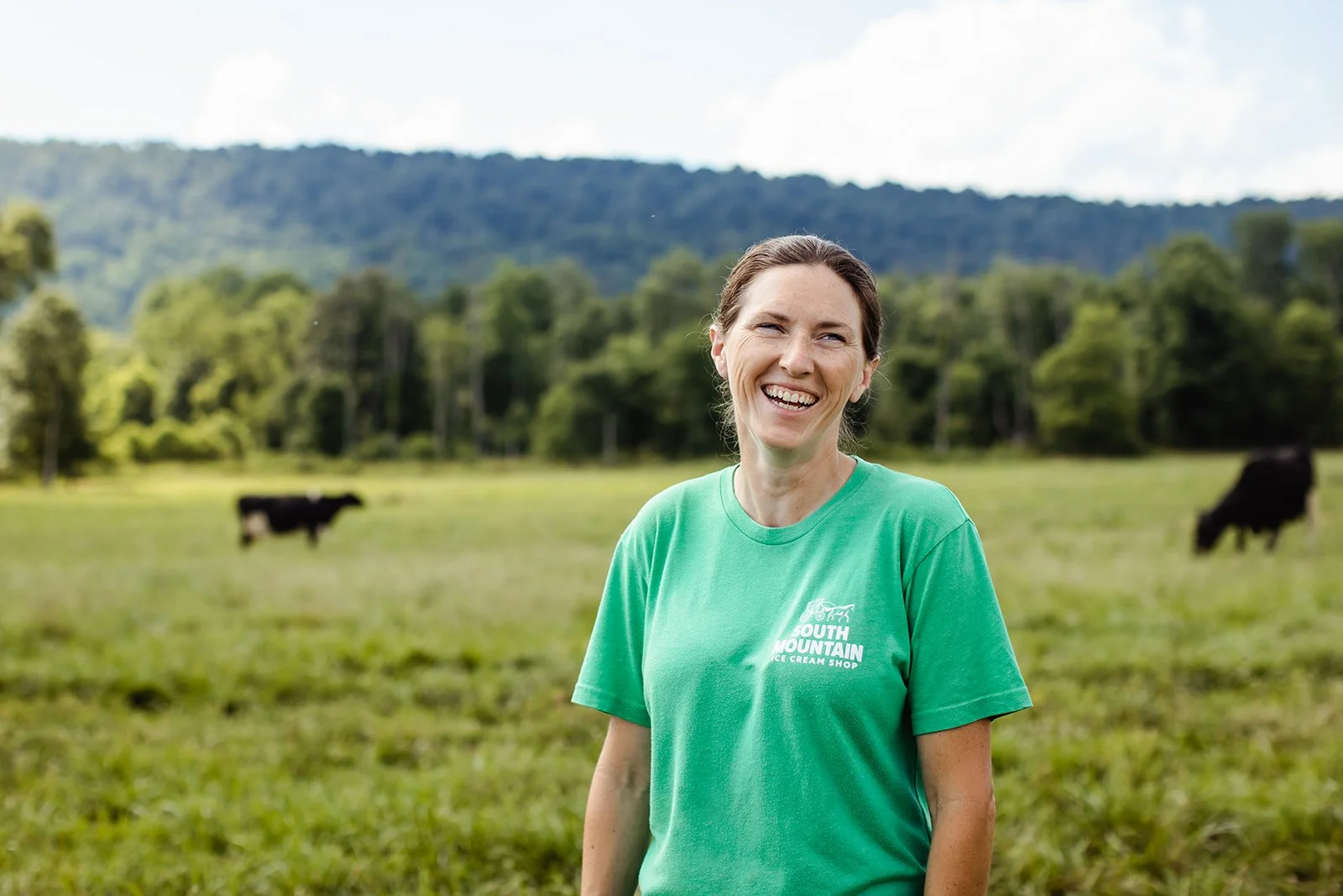 A woman smiling in a green South Mountain Ice Cream Shop t-shirt stands in a grassy field with cows grazing and a mountain in the background.