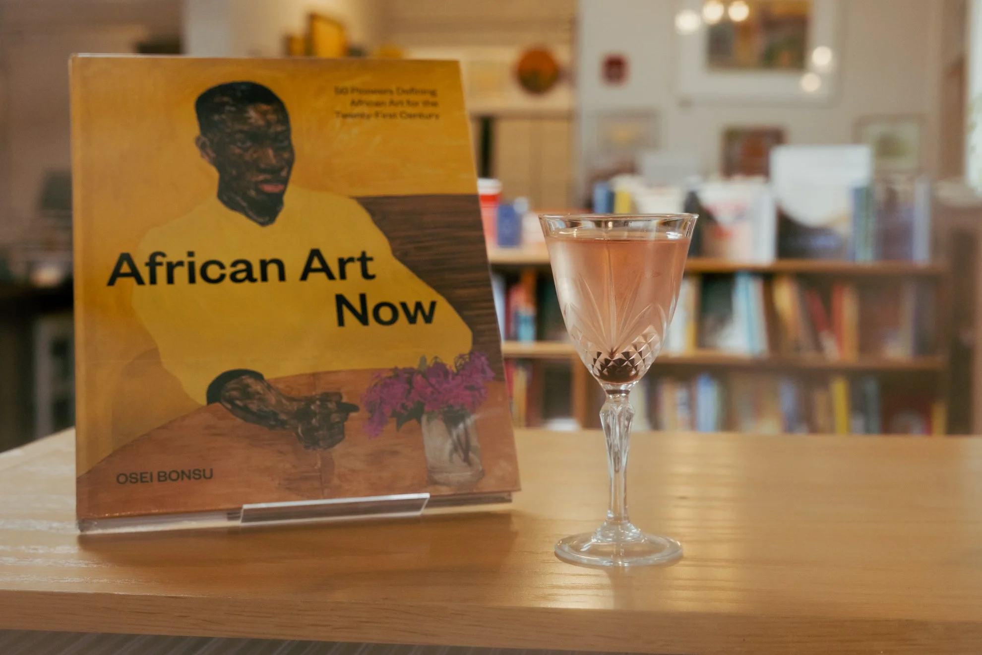 A magazine titled 'African Art Now' on a stand, with a glass of pinkish drink on a wooden table, in a room with many bookshelves in the background.