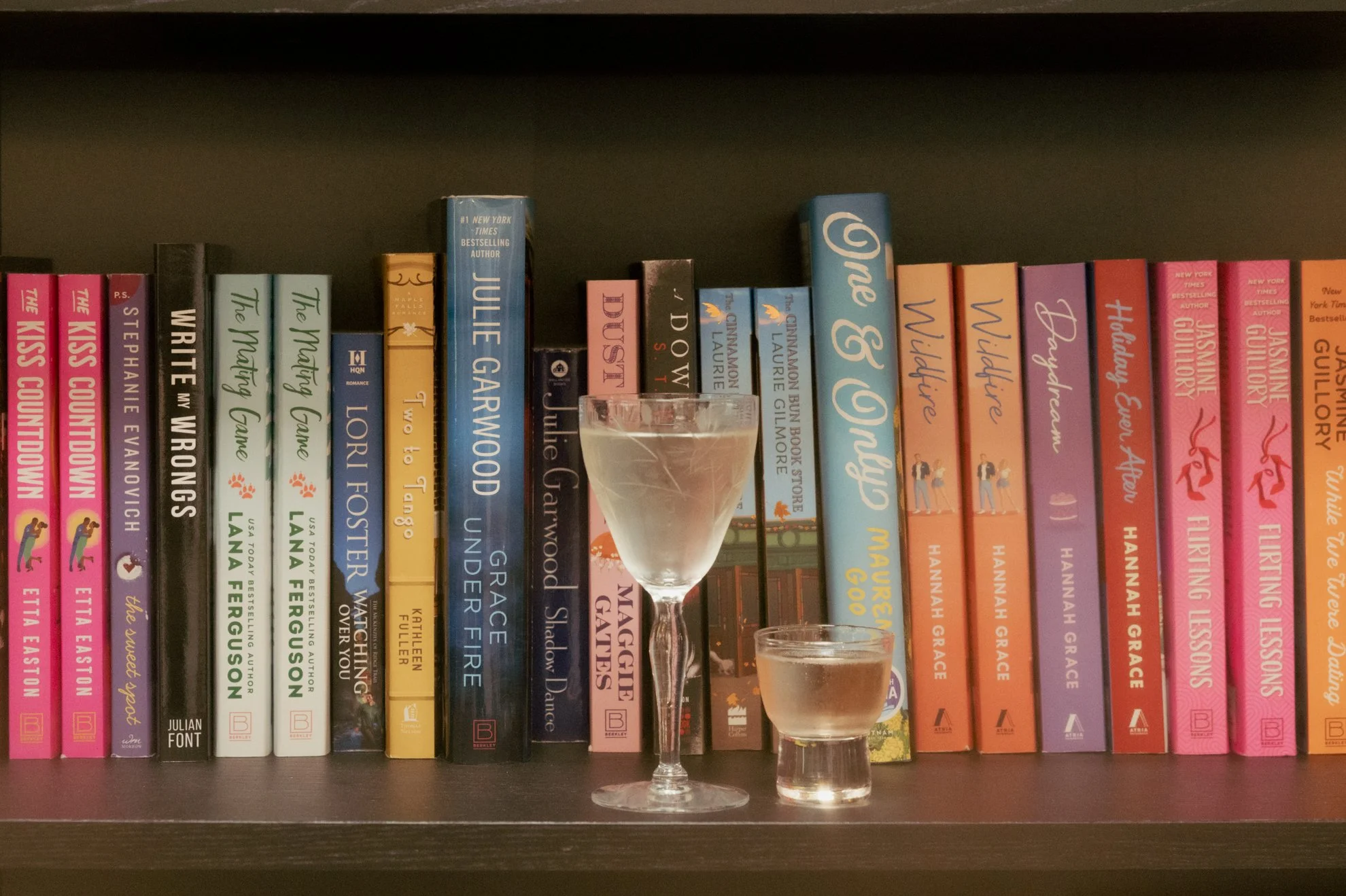 A bookshelf filled with colorful books, with two shot glasses in the foreground, one containing a clear liquid and the other an amber liquid.