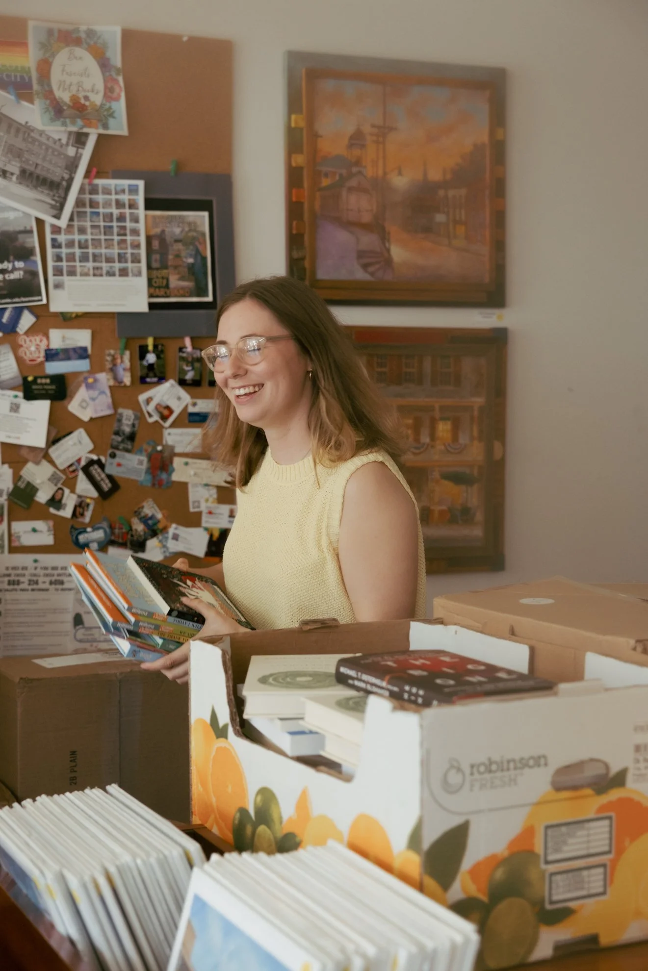 A woman with glasses smiling and holding a book in a store with books and artwork on the wall behind her.