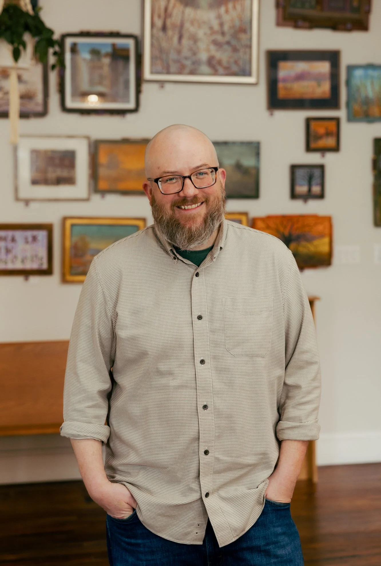 A smiling man with glasses, a beard, and a shaved head standing in an art gallery with framed paintings on the wall behind him. He is wearing a light-colored checkered shirt with the sleeves rolled up and blue jeans.