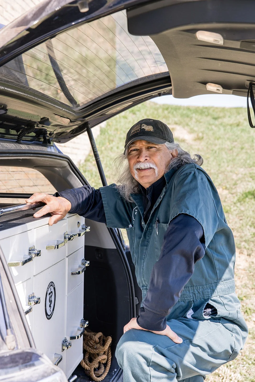 An older man with long gray hair and a mustache is kneeling next to an open vehicle trunk, smiling at the camera. He is wearing a cap, a dark jacket, and gray pants. Inside the trunk is a white medical or emergency cabinet with multiple drawers and s