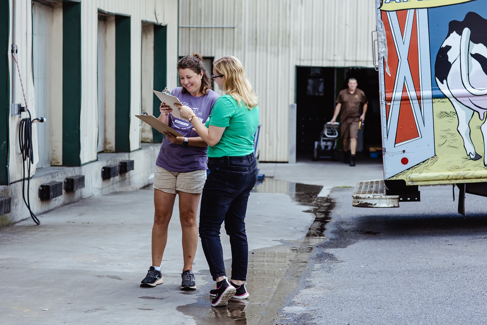 Two women standing outside, looking at a clipboard and smiling, with a mass of water on the ground nearby. In the background, a man is walking towards a colorful trailer.