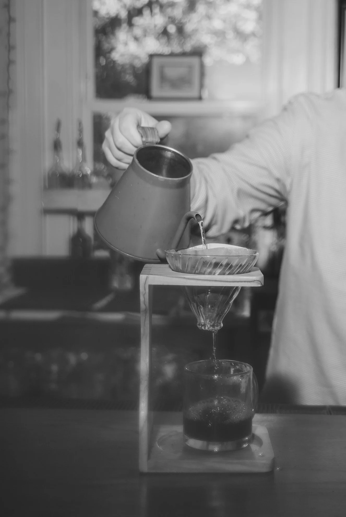 A person in a long-sleeved shirt pours a beverage into a glass through a funnel. The setup appears to be a home or studio environment with a window in the background and outdoor scenery visible. The image is black and white.