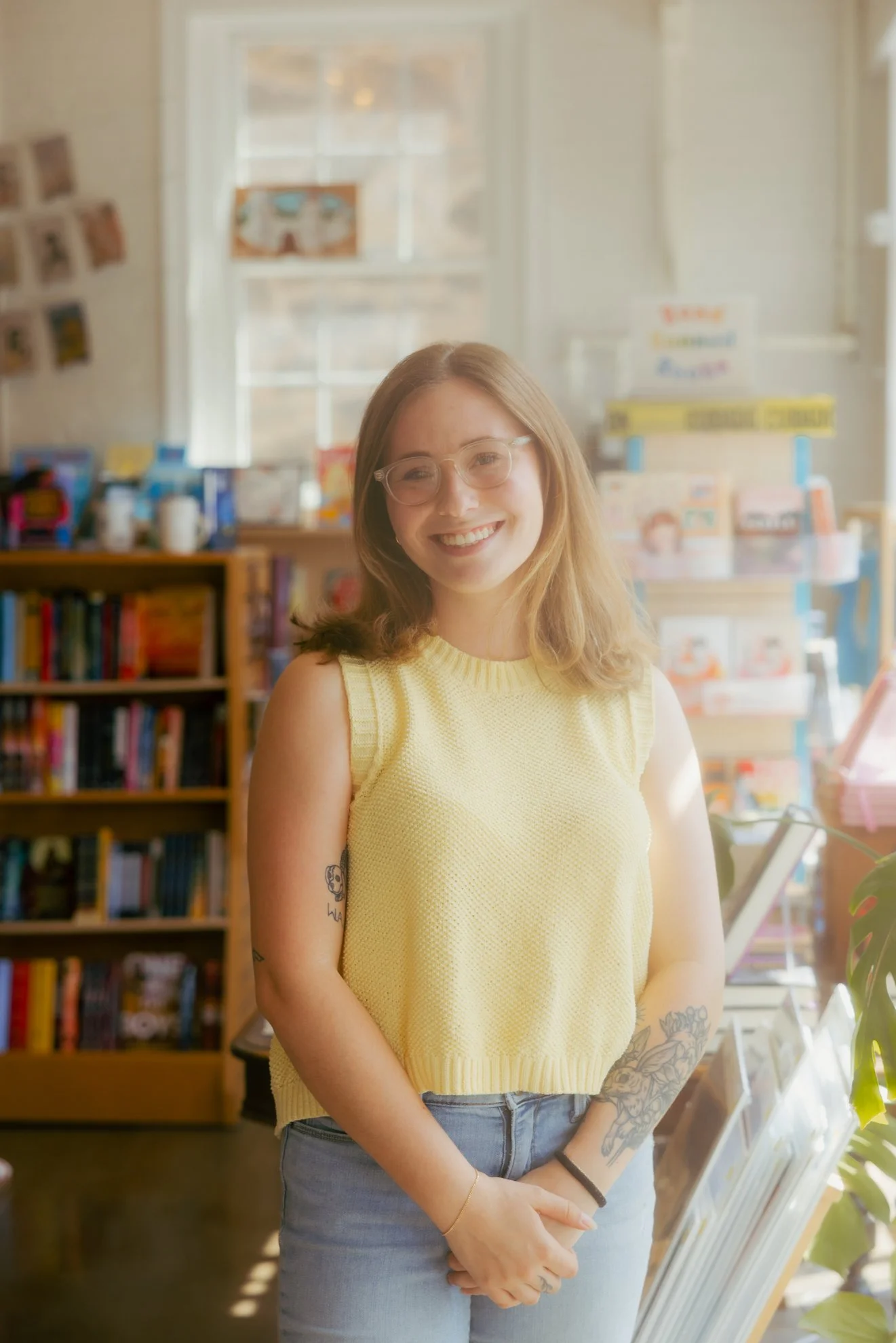 A woman with glasses and tattoos smiling in a bookstore or library, wearing a yellow sleeveless top and blue jeans, with shelves of books and bright sunlight in the background.