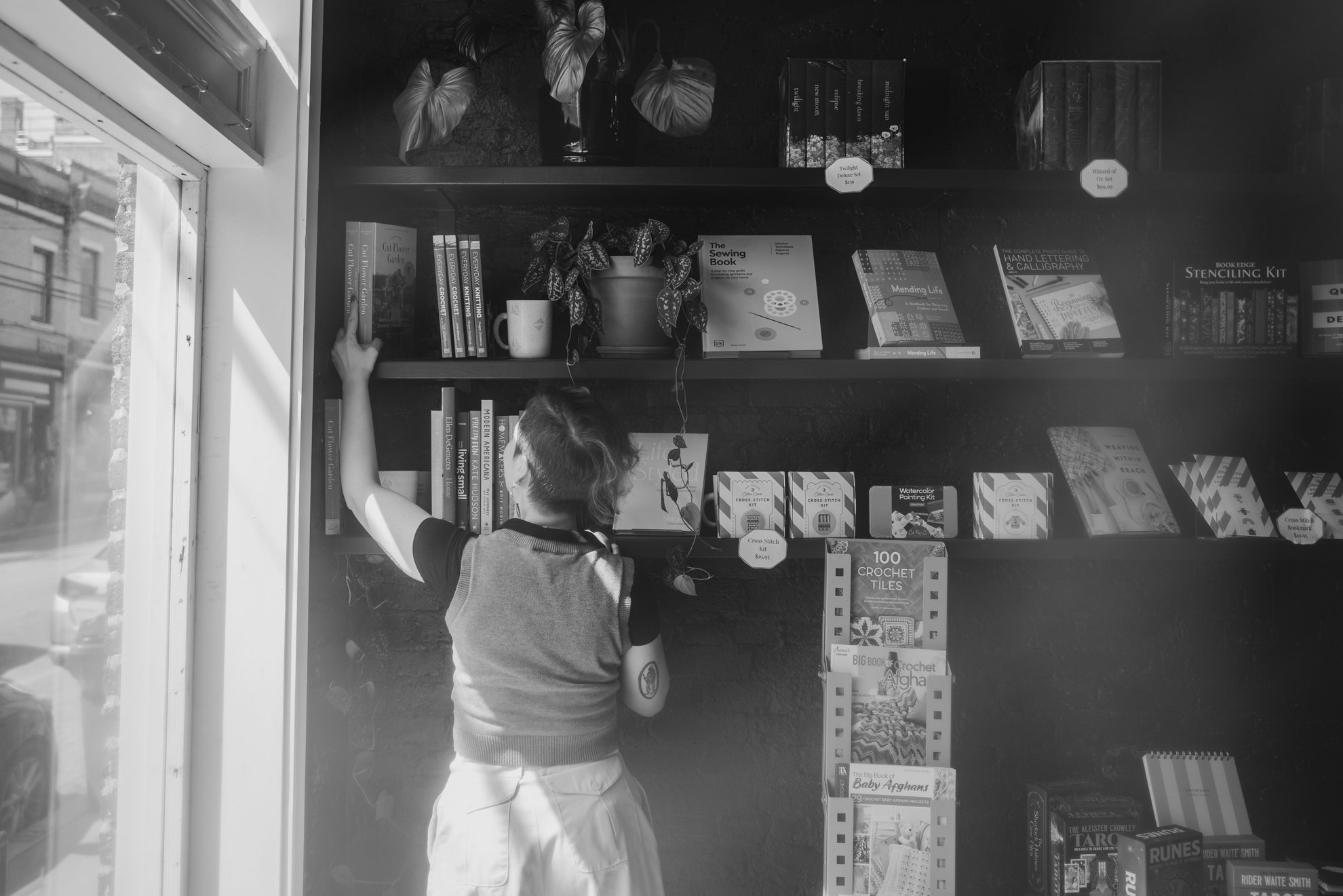 A young boy with short hair, wearing a striped shirt and white shorts, is reaching for a book on a bookshelf inside a store. The bookshelf contains various books and decorative items, and there is sunlight streaming in from a nearby window on the lef
