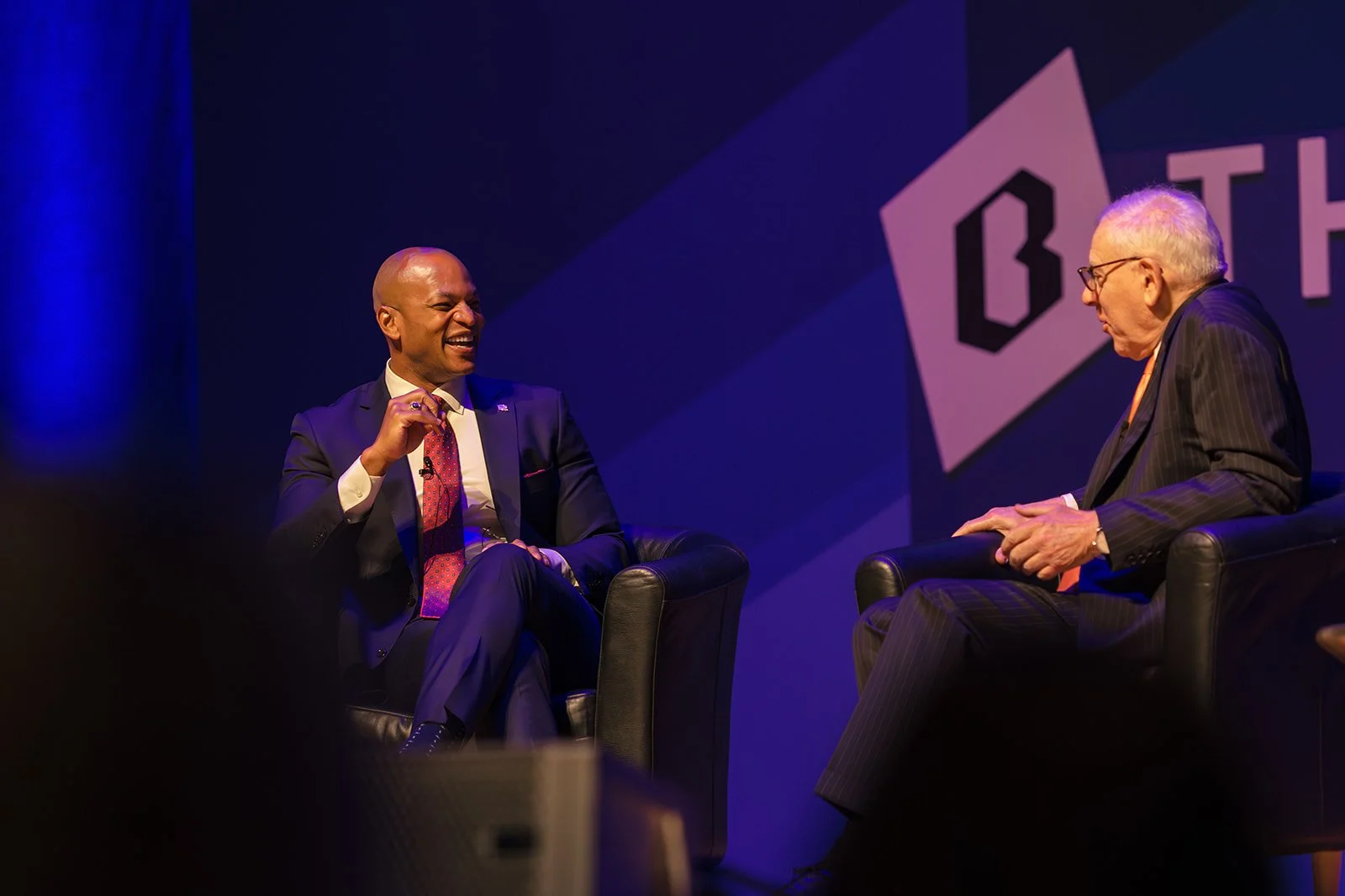 Two men in suits sitting in armchairs on stage, engaged in conversation during a panel discussion, with a large logo and the word 'BATCH' in the background.