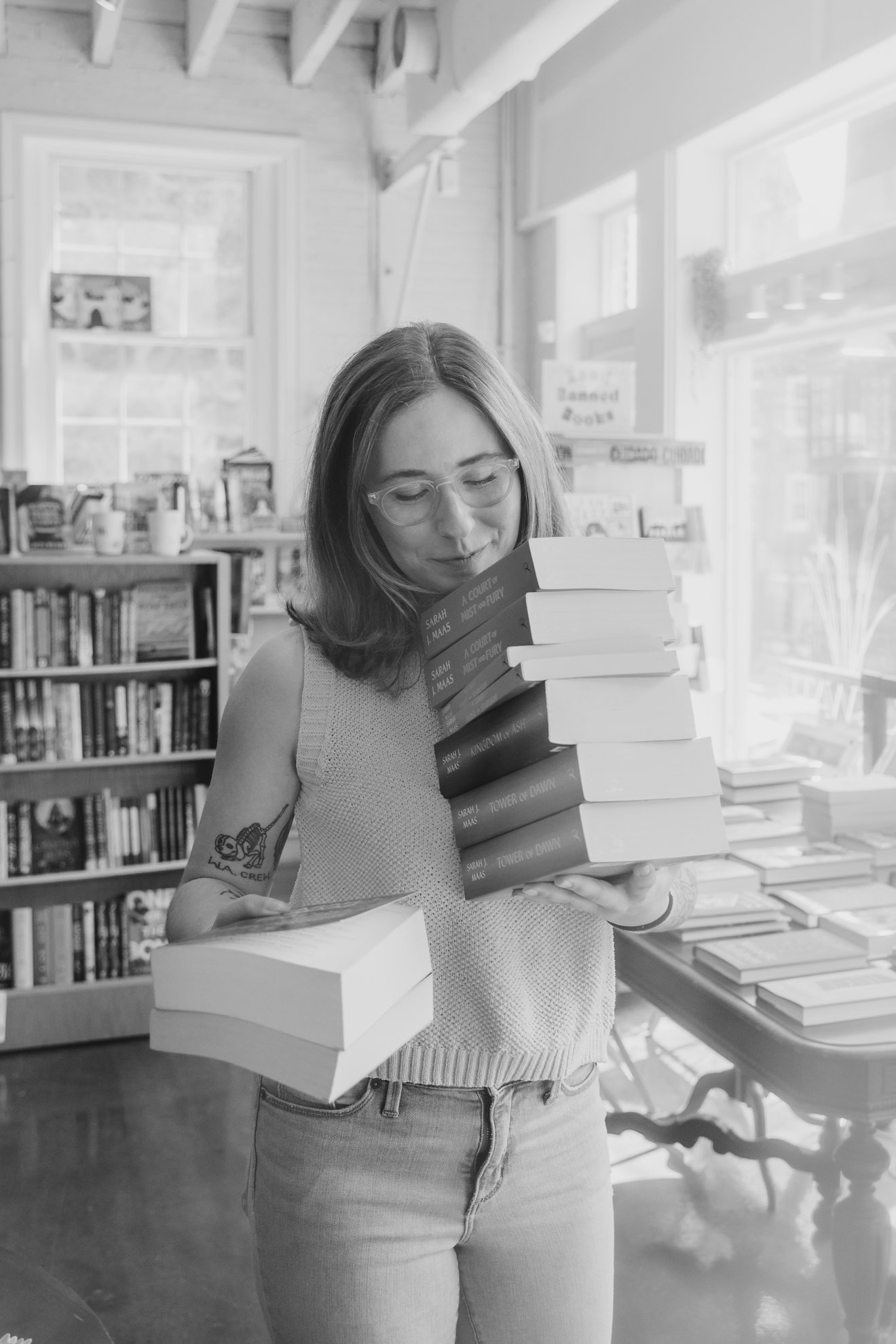 A woman with glasses and shoulder-length hair stands inside a bookstore, holding a stack of books and looking down at them. She has a tattoo on her left arm and is wearing a sleeveless top and jeans.