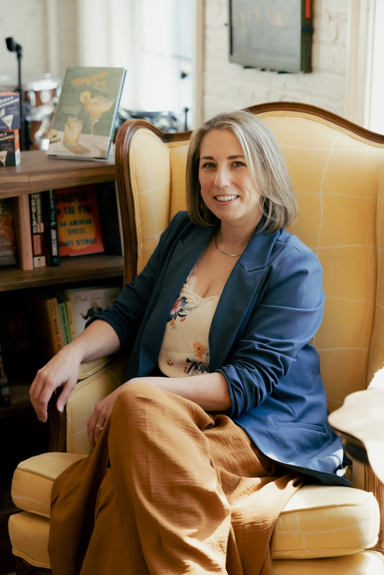 A woman with shoulder-length gray hair wearing a blue blazer and floral blouse, sitting in a yellow armchair in a cozy room with bookshelves and a television in the background.
