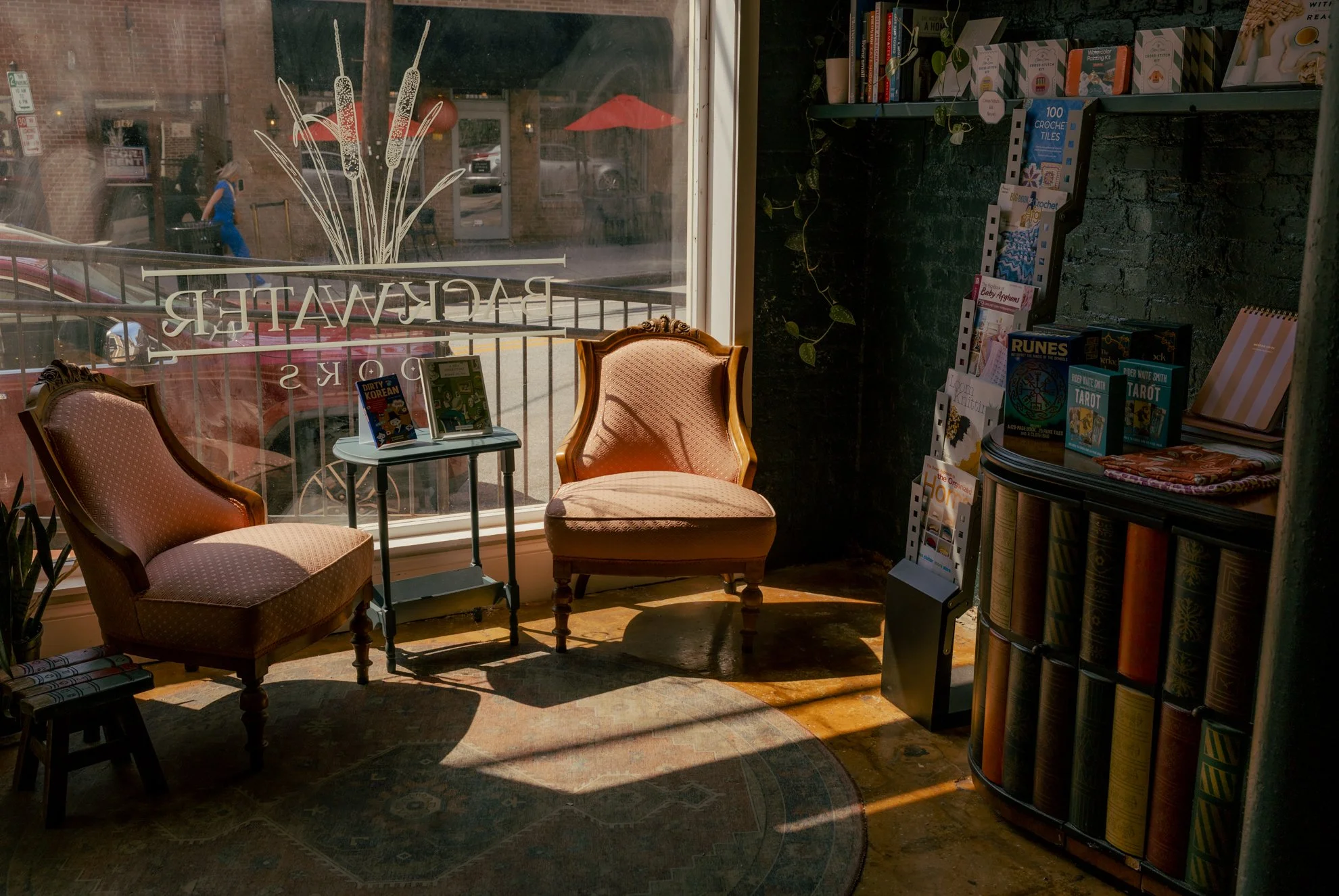 A cozy bookstore or coffee shop interior with two vintage armchairs near a large window. The window has a decal of a plant and reverse text 'RAREWATER.' Outside, there is a sidewalk and a person in a blue outfit walking past brick buildings. Inside, 