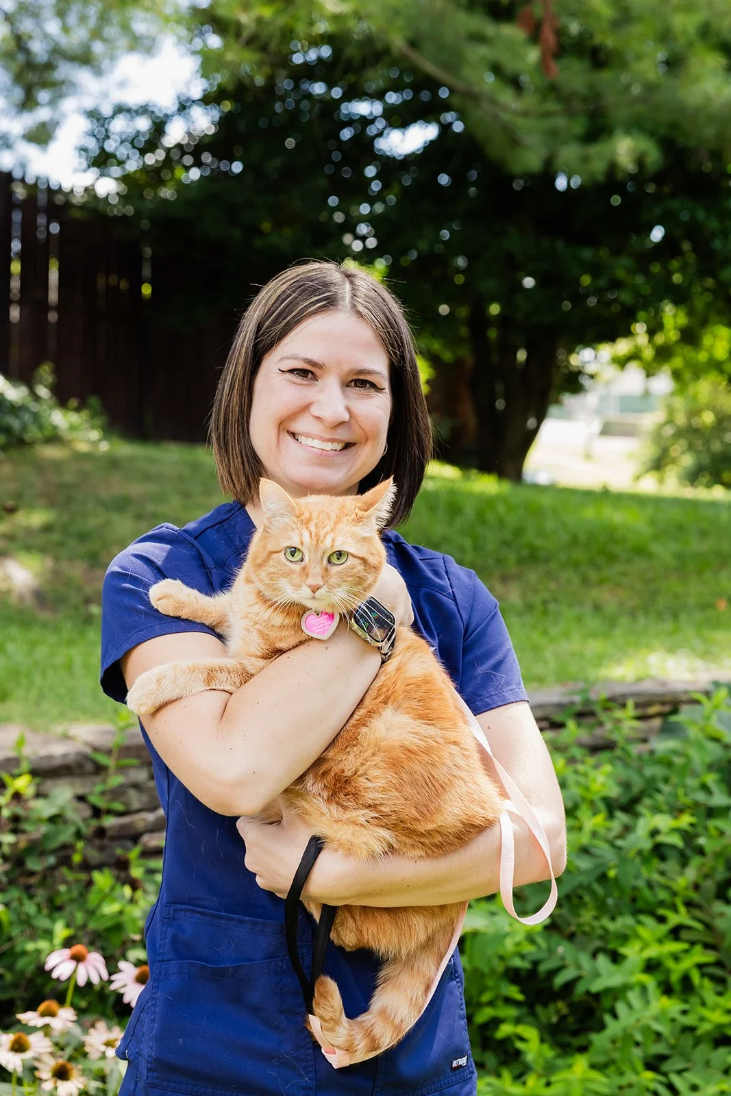 A woman with short brown hair smiling and holding an orange tabby cat outdoors in a garden with green trees and flowers in the background.