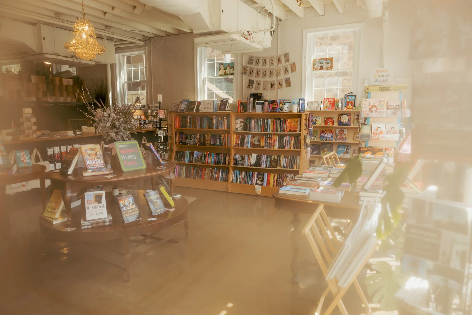 A cozy bookstore with several bookshelves full of colorful books, a circular display table showcasing recent releases, large windows letting in natural light, decorated with a banner that says 'Once Upon a Time,' and a chandelier hanging from the cei