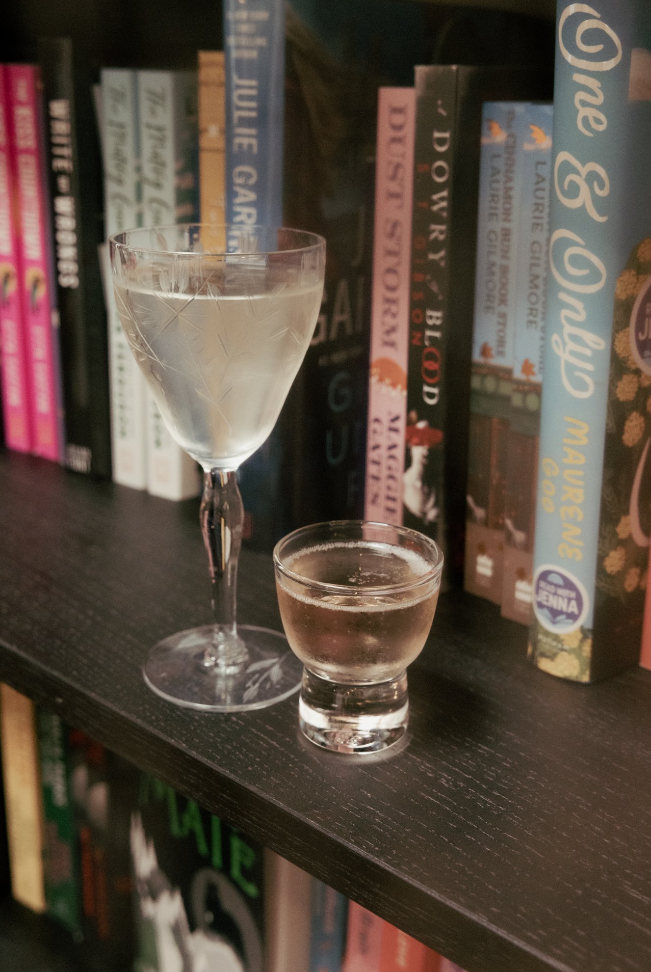 A close-up photo of two drinks, one cocktail in a tall glass with a stem and a smaller shot glass, placed on a black shelf in front of a row of colorful books.