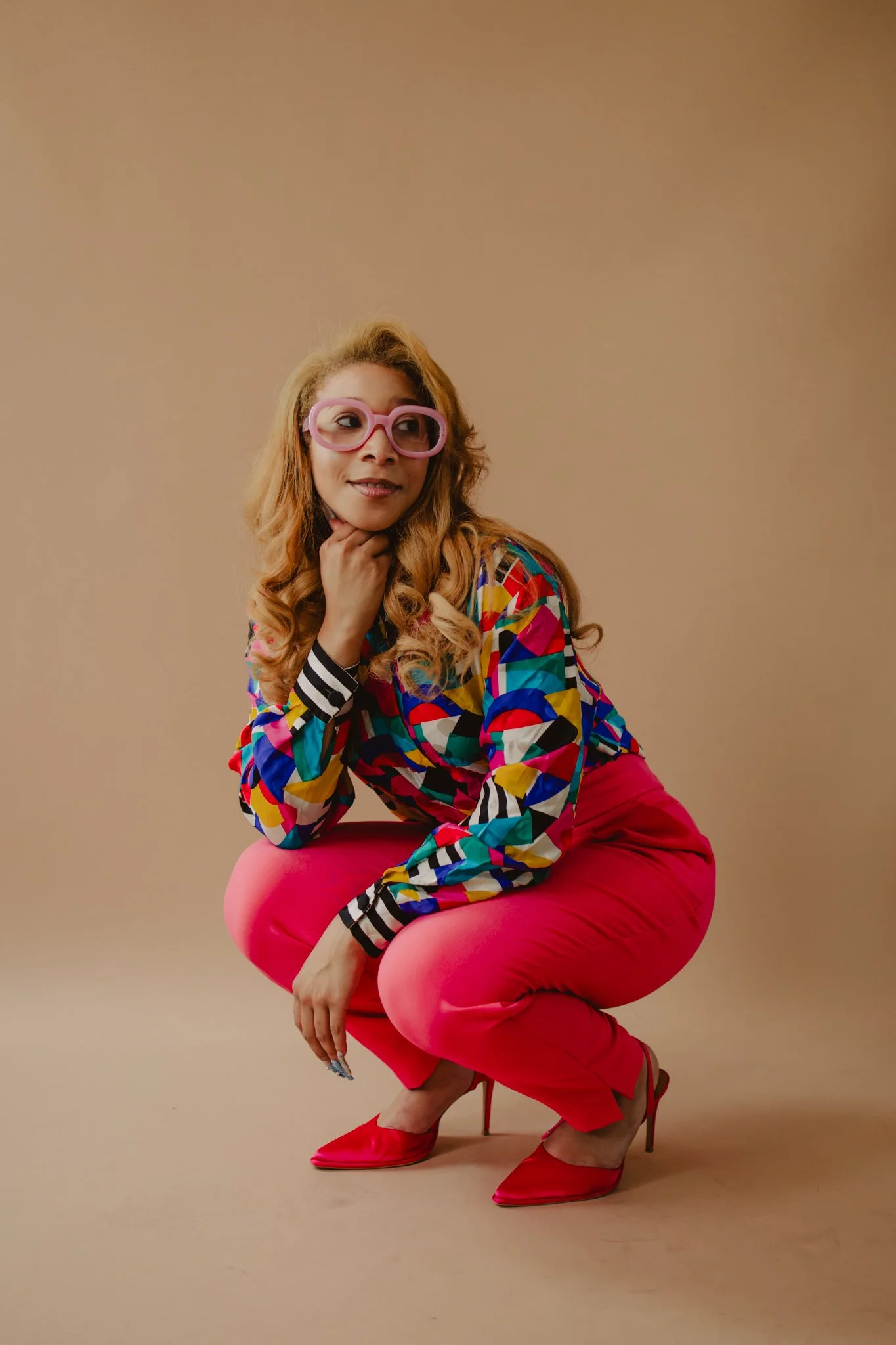 A woman with wavy blonde hair wearing large pink glasses, a colorful geometric-patterned blouse, red pants, and red high heels, sitting on a pink stool against a neutral background.